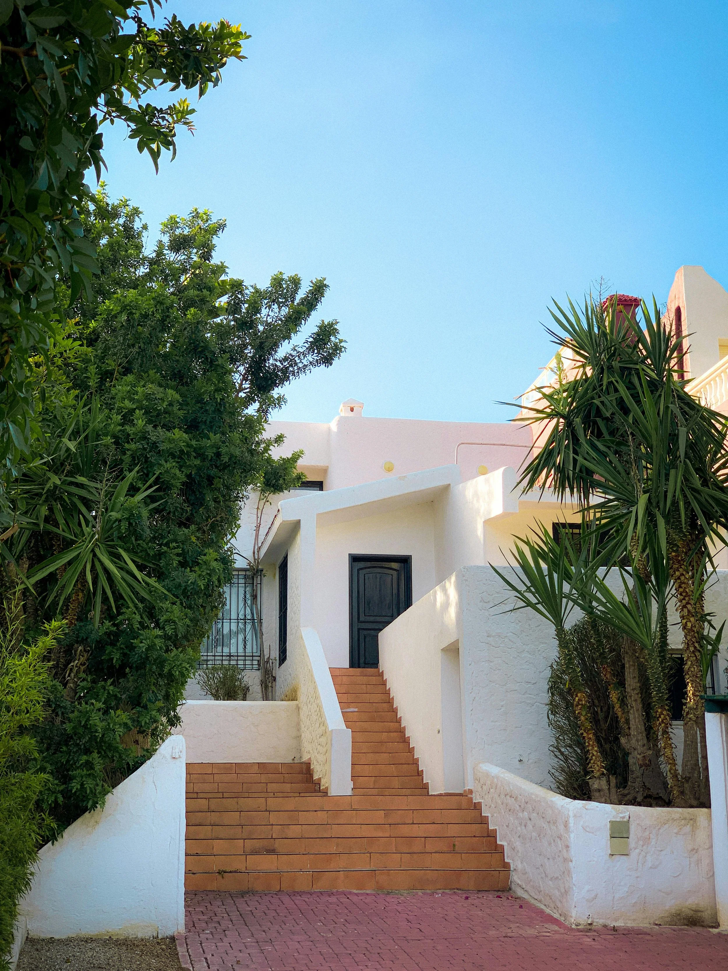 White Mediterranean-style house with stairs, surrounded by green trees and plants, under a clear blue sky.