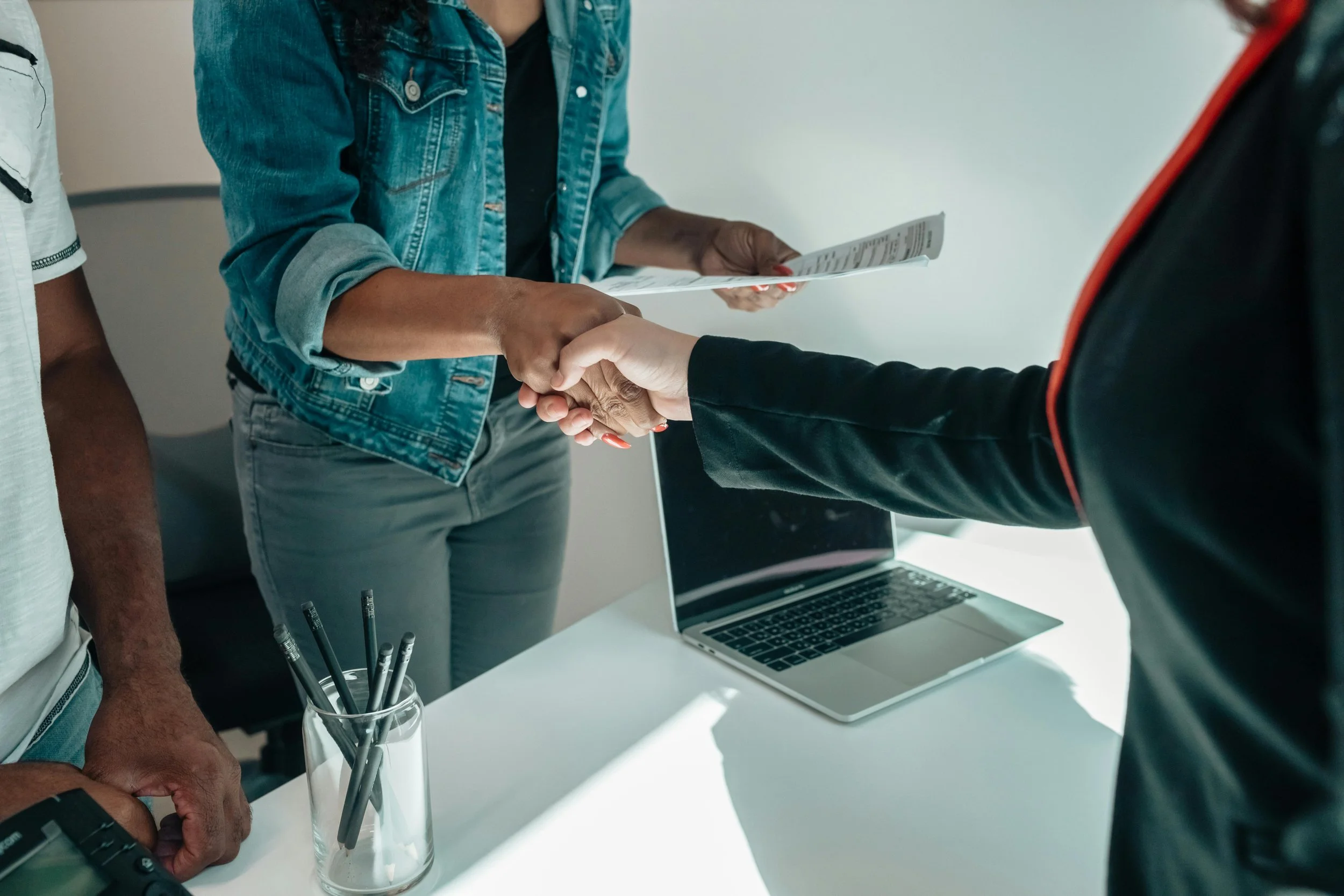 People shaking hands in an office, with documents, a laptop, and a glass containing pens on the table.