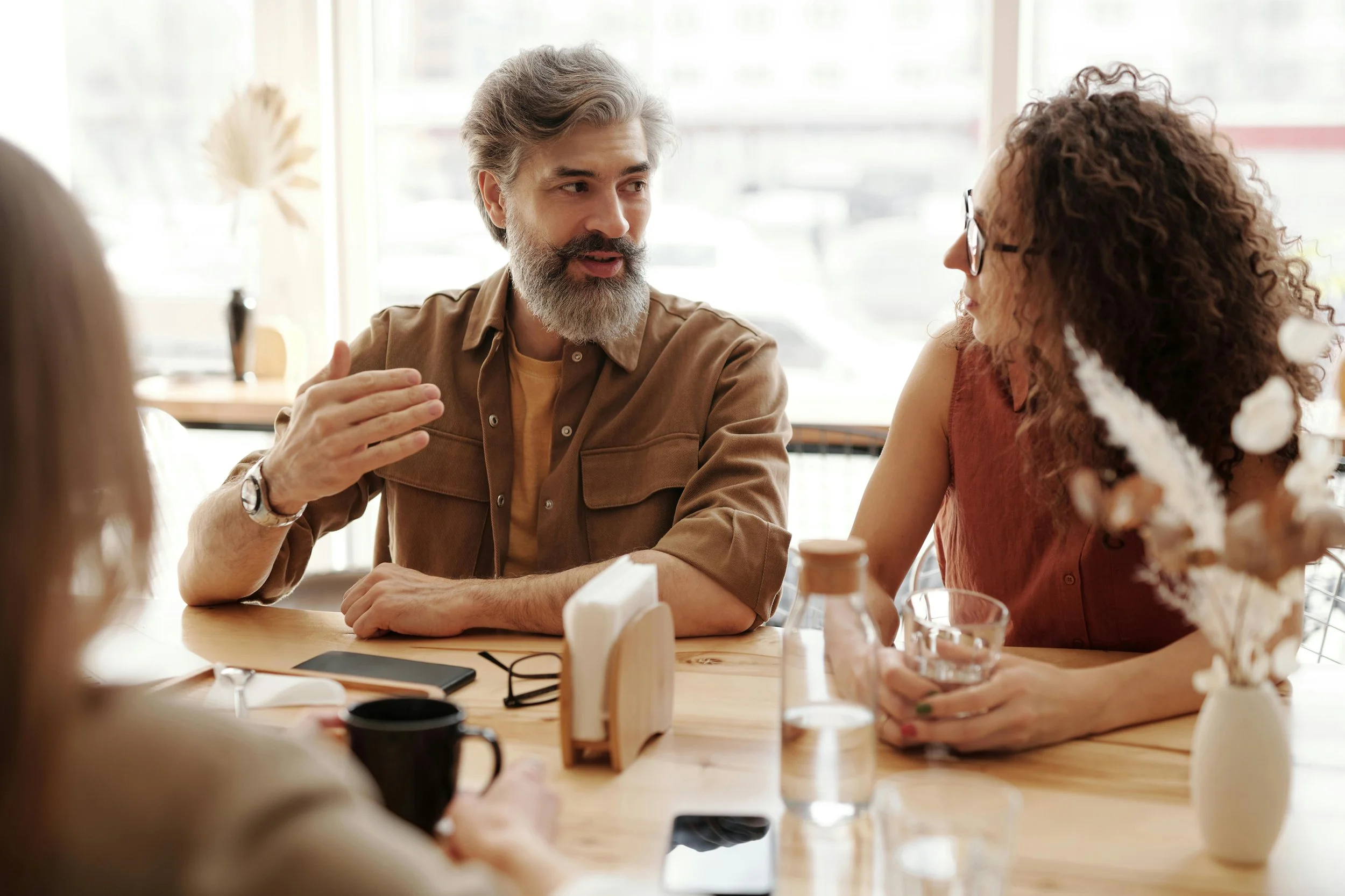 A middle-aged man with a beard and gray hair engaged in conversation with a curly-haired woman wearing glasses at a dining table in a well-lit café.