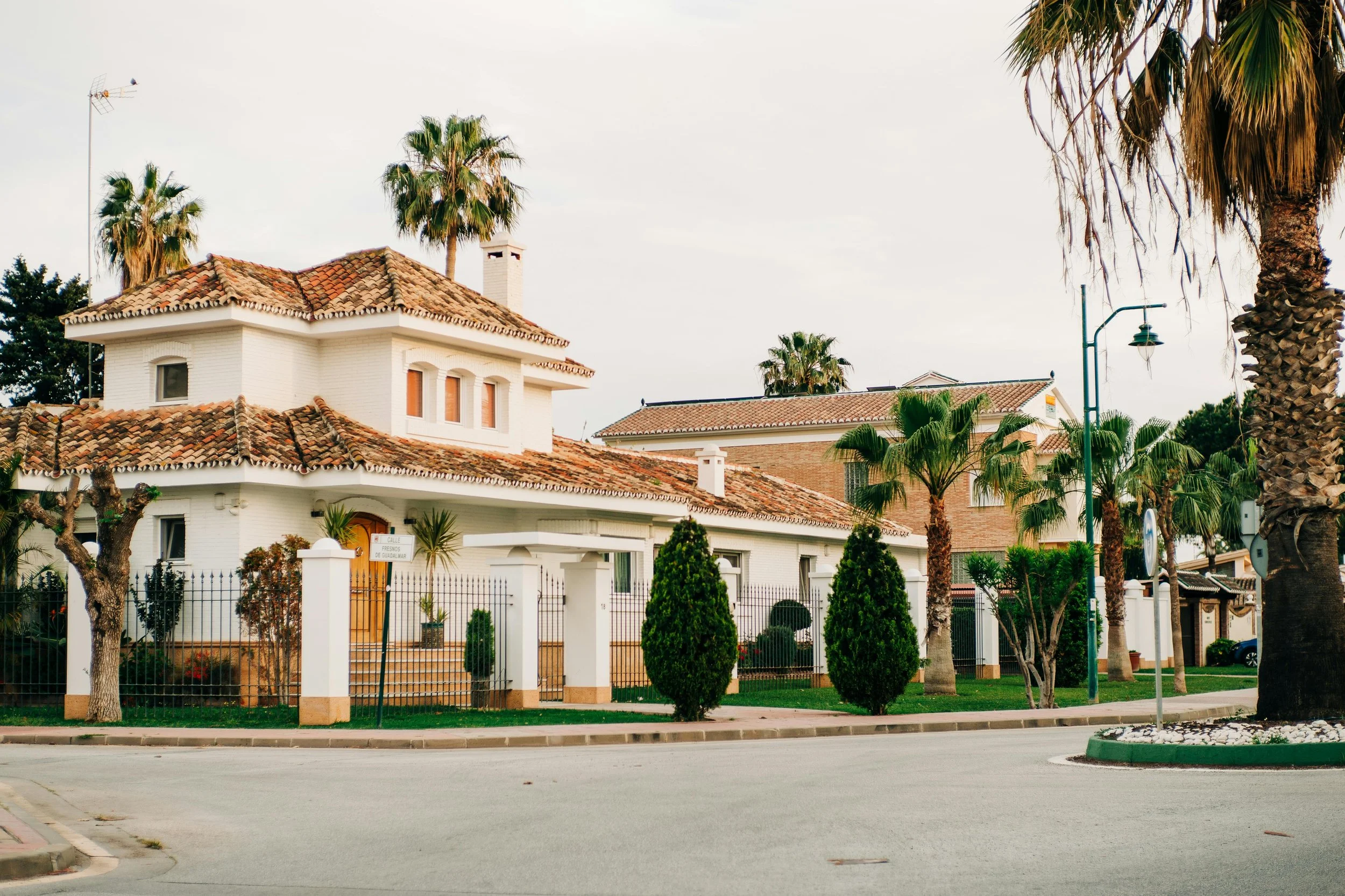 Residential neighborhood with white houses, tiled roofs, and tall palm trees along the street under a cloudy sky.