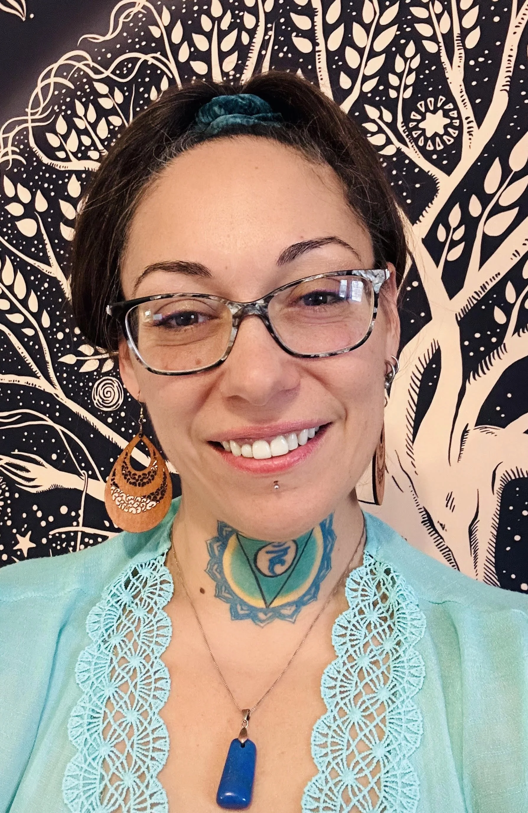 A woman with glasses smiling in front of a black and white patterned background. She has dark hair with a teal headband, large earrings, a tattoo on her chest, and is wearing a turquoise top with lace detailing.