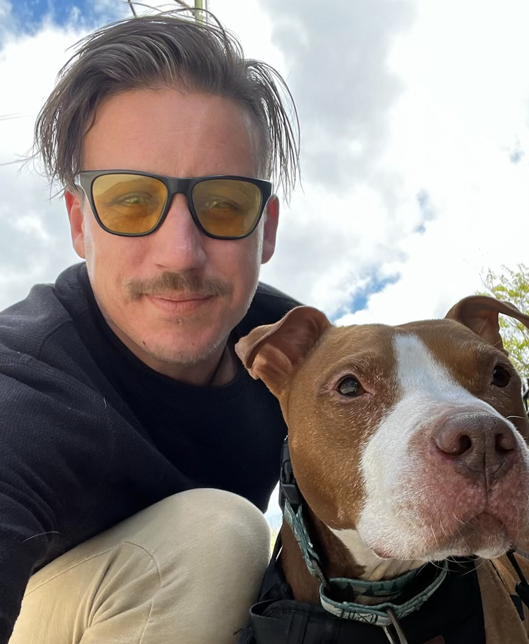 A man wearing sunglasses with yellow lenses takes a selfie with his dog outdoors on a cloudy day.
