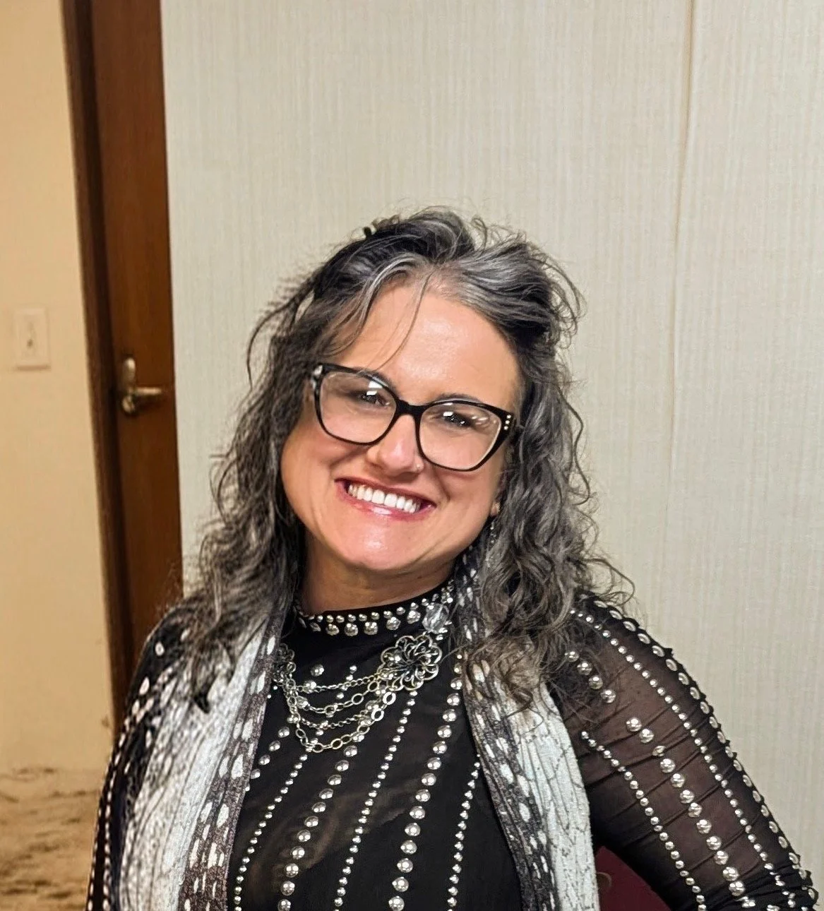 A smiling woman with gray curly hair, black glasses, wearing a black top with pearl embellishments, and layered silver necklaces, standing indoors against a beige wall.