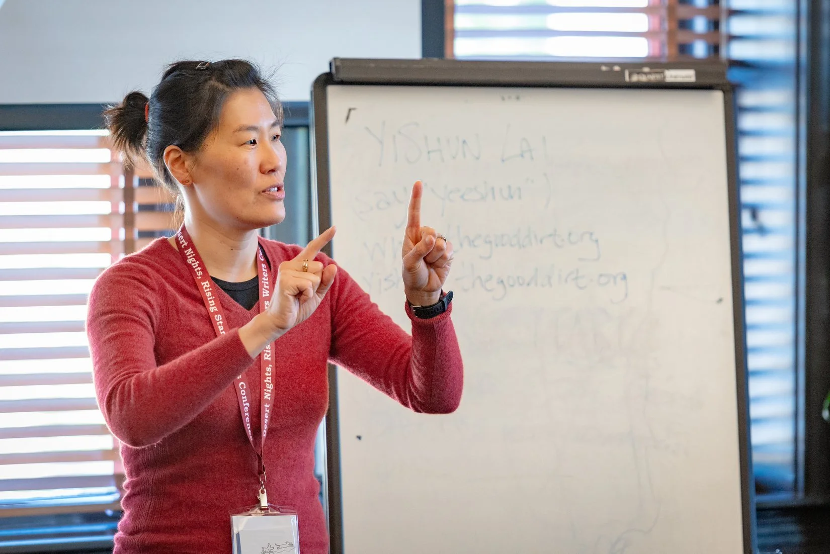 A woman with dark hair pulled back, wearing a red sweater and a conference badge, is speaking in front of a whiteboard with handwritten notes. She is using hand gestures to emphasize her point.