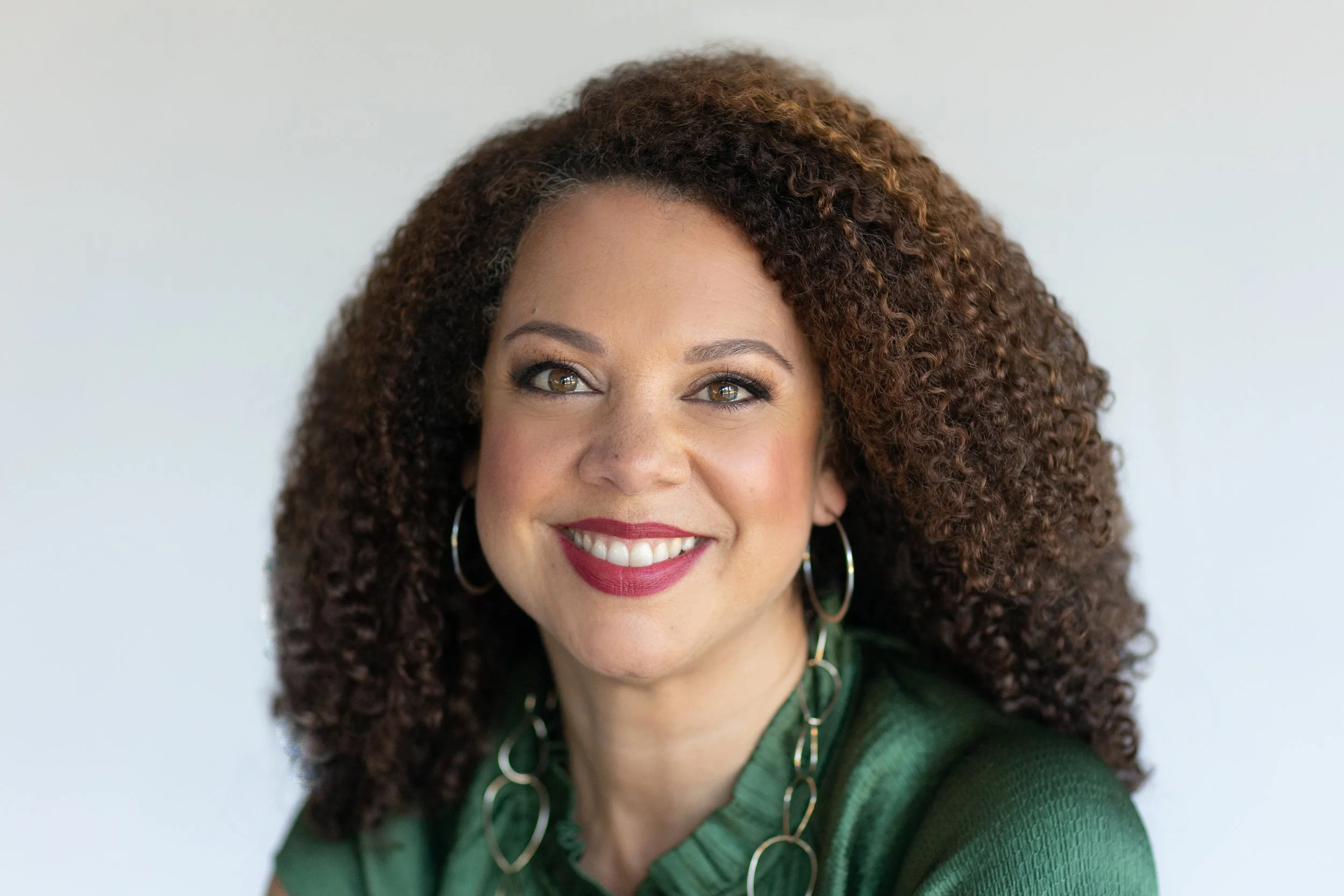 Close-up portrait of a smiling woman with curly brown hair, wearing hoop earrings and a green top.