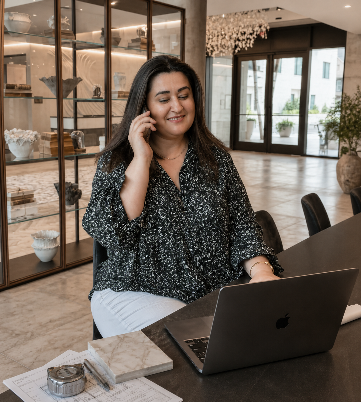 A woman sitting at a table in a modern, well-lit lobby or reception area, talking on a cellphone and working on a laptop.