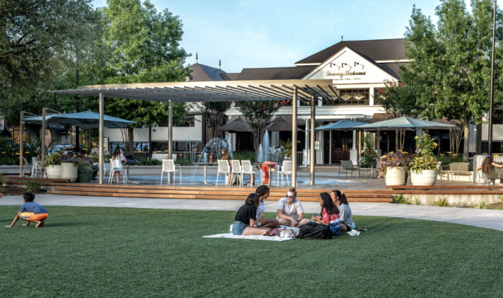 People sitting on a grassy area in front of a shopping center with a fountain, trees, and outdoor seating.