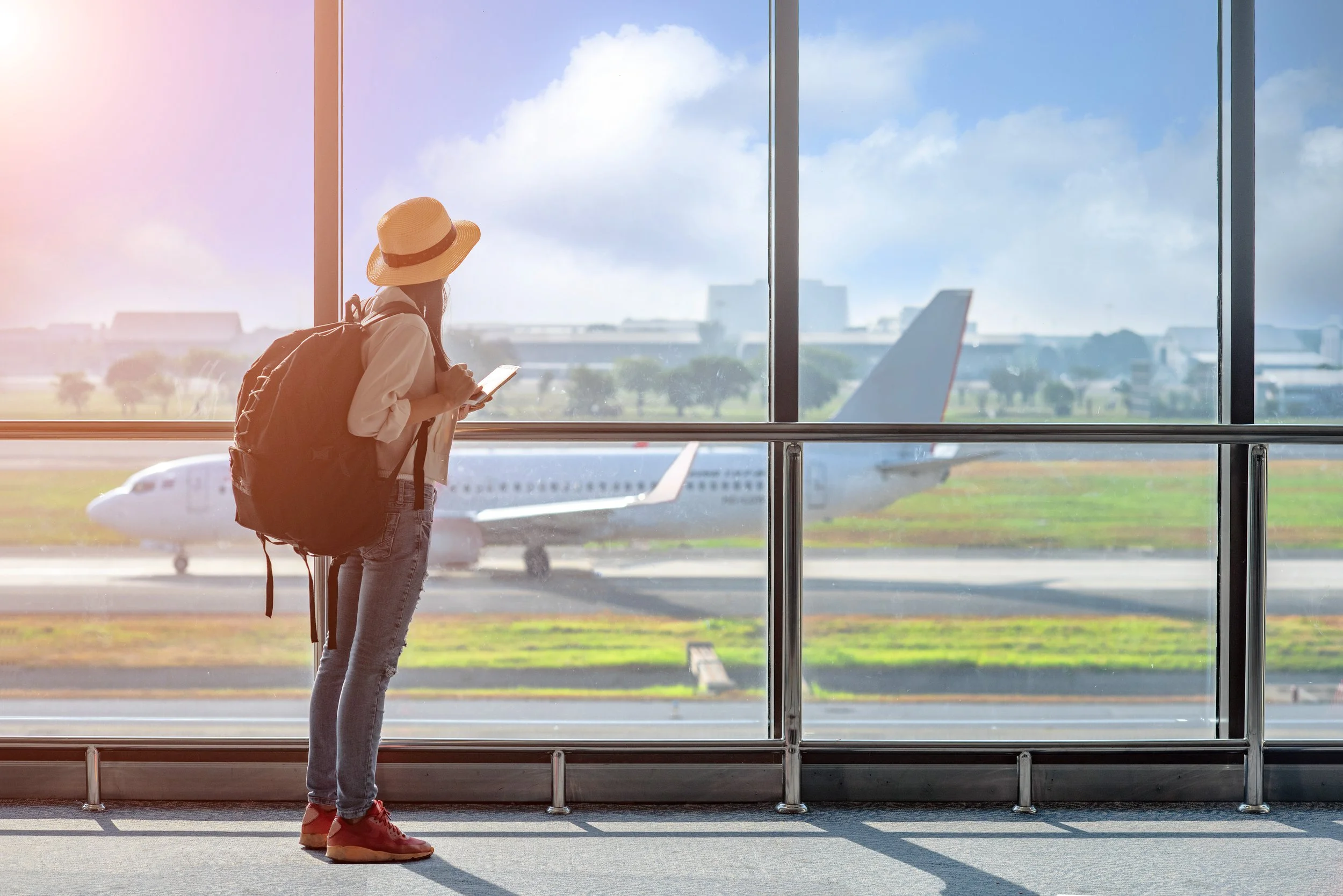 A woman with a backpack, wearing a wide-brimmed hat and pink sneakers, standing at an airport terminal window, looking out at the runway and airplanes outside on a sunny day.