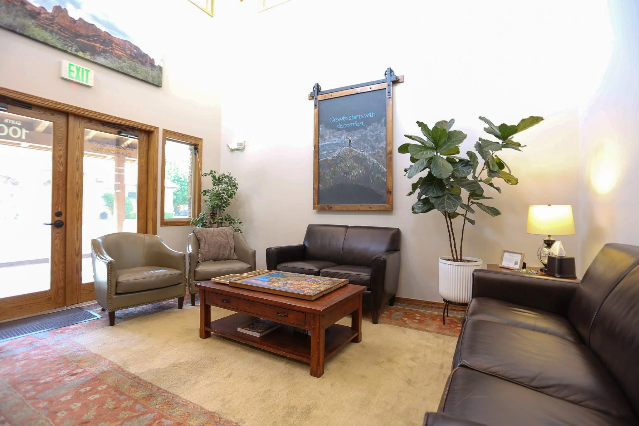 Wind Rivers Counseling waiting room with beige and brown couches, a wooden coffee table, a large potted plant, and a lamp. There are windows and doors with wooden frames, and a chalkboard with a quote on the wall.