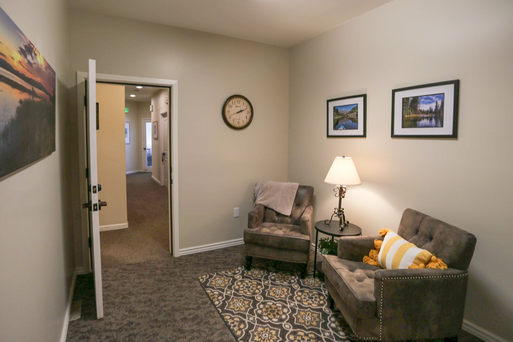 Sitting area near therapist offices, a grey armchair with a throw blanket, a small round side table with a lamp, framed landscape photos on the wall, and a patterned rug.