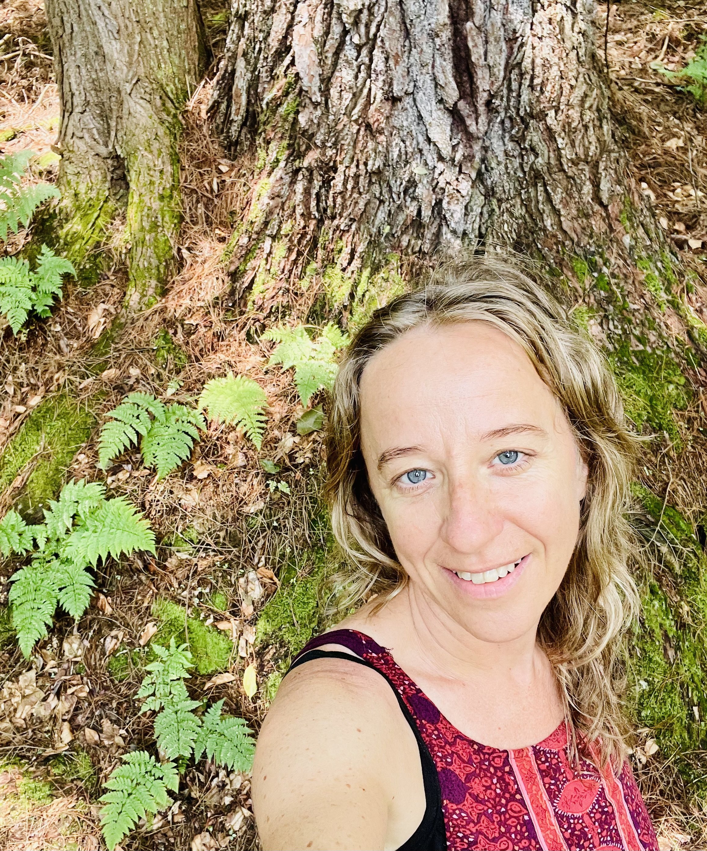Woman with blue eyes and curly blonde hair lying on forest ground, smiling, near a large tree trunk surrounded by green plants and fallen leaves.