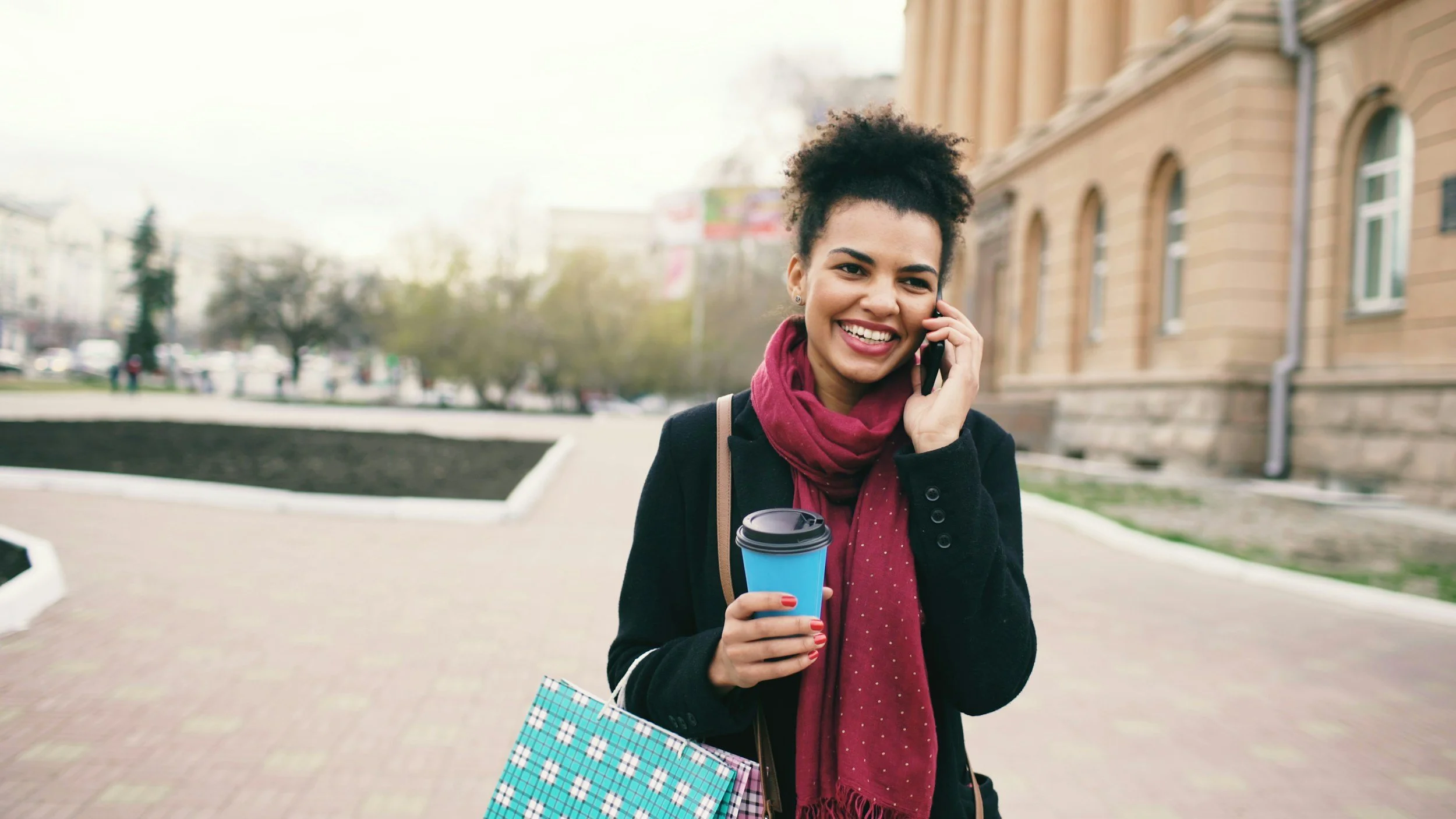 Smiling young woman talking on cellphone outdoors near a building, holding a coffee cup, carrying shopping bags.
