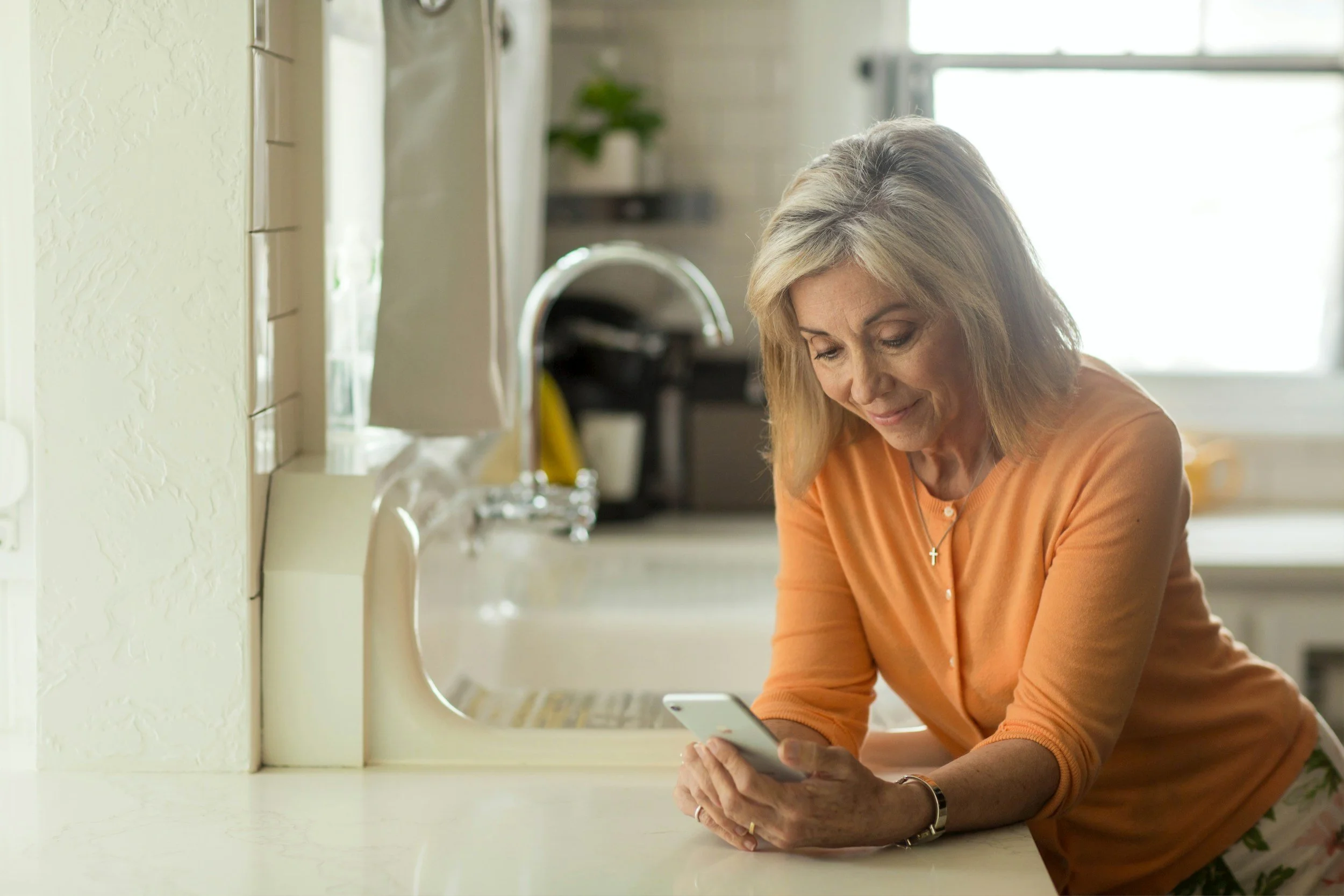 Woman in orange top using a smartphone in a kitchen.