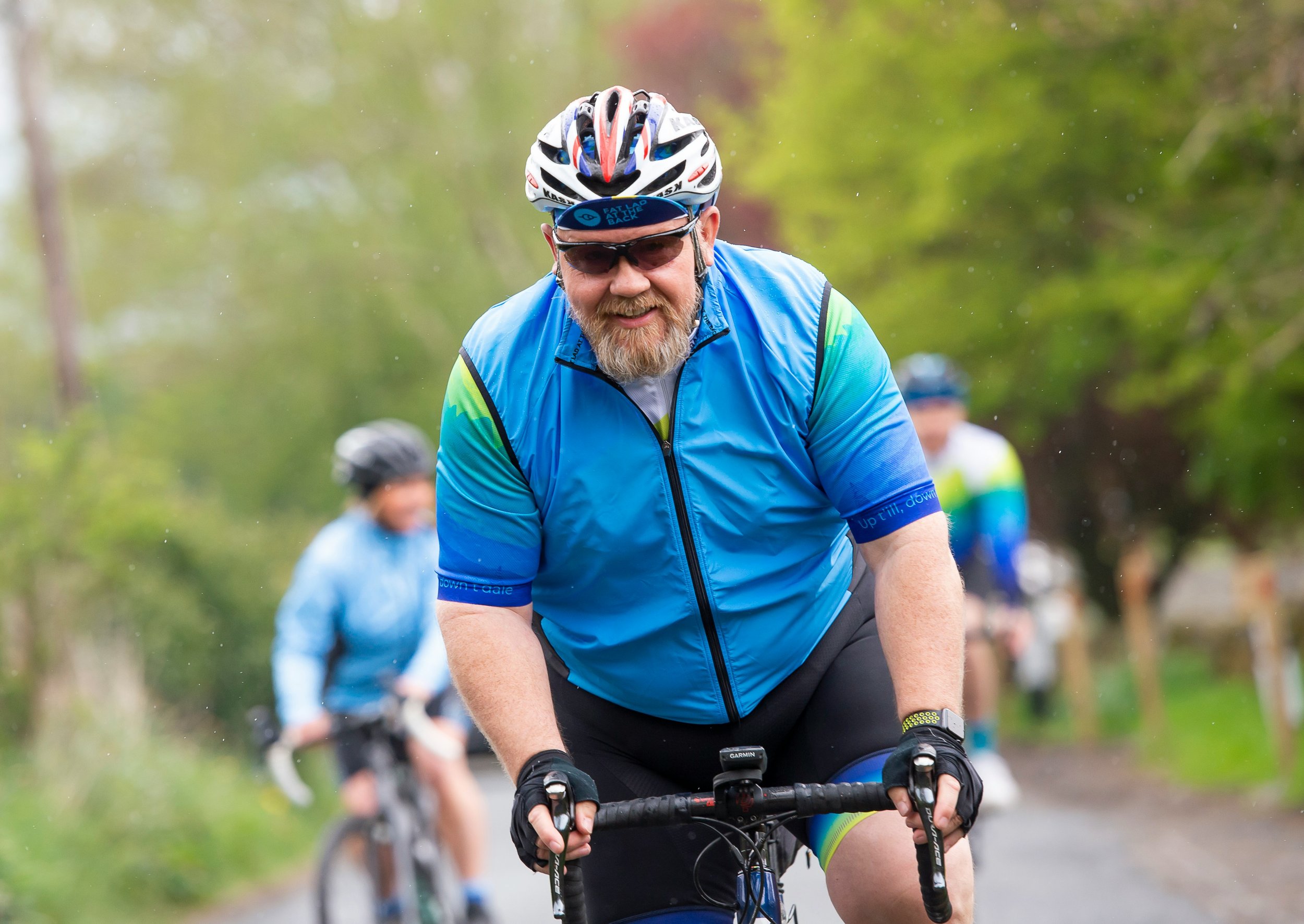A man with a beard and sunglasses riding a mountain bike outdoors, wearing a blue cycling shirt, helmet, and gloves, with other cyclists in the background on a trail surrounded by trees.