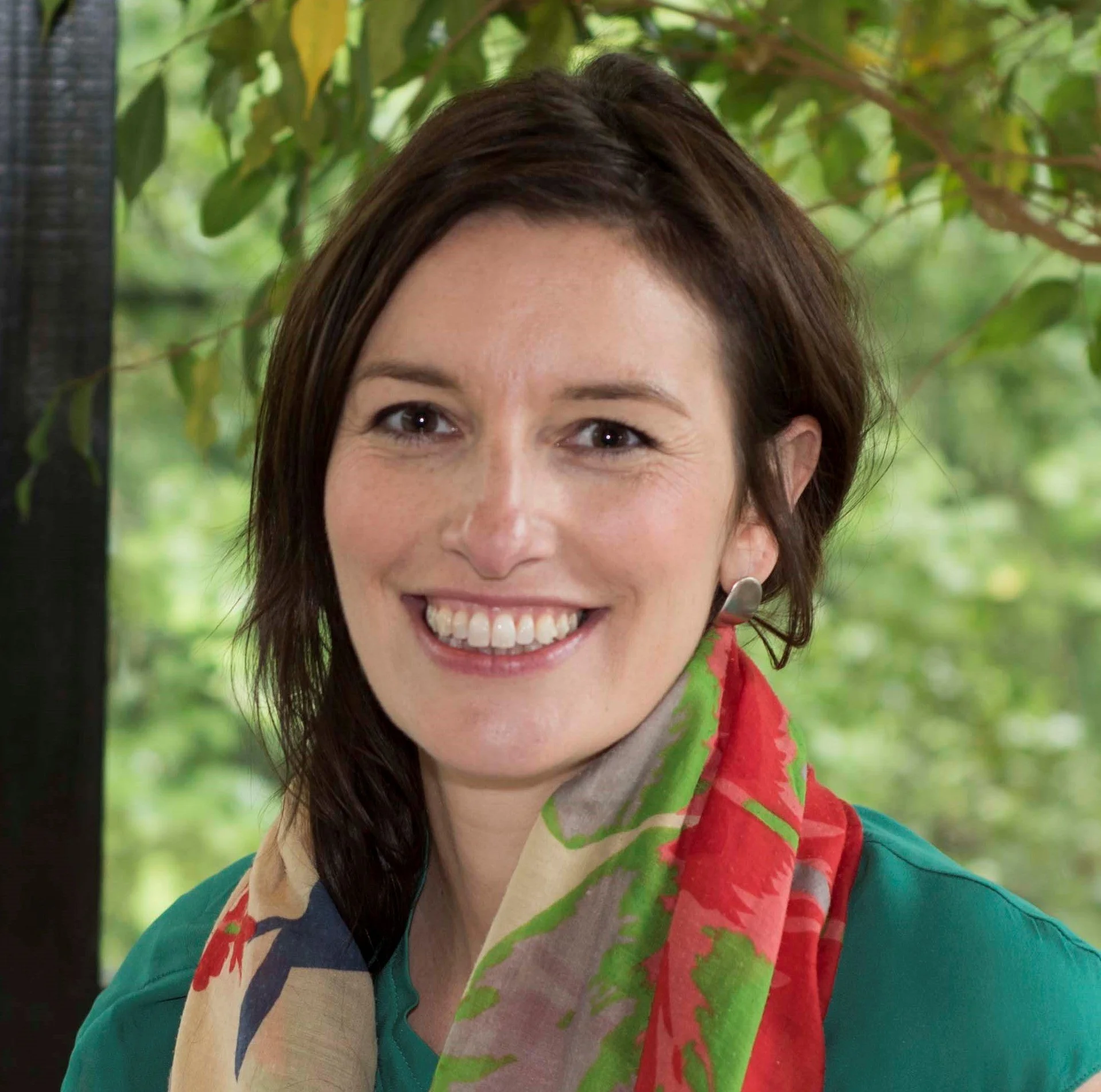 Smiling woman with short brown hair, wearing a colorful scarf and a teal shirt, standing outdoors with green foliage in the background.