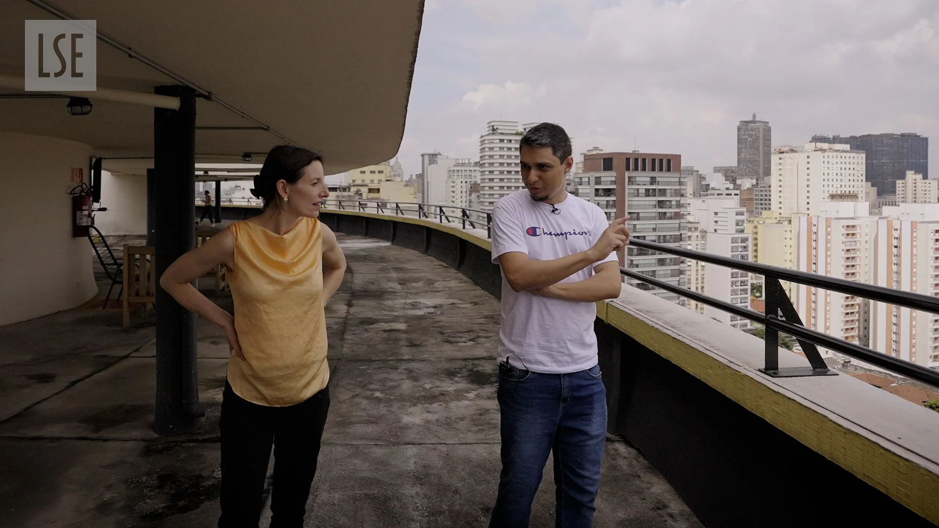 Two people, a woman and a man, are standing on a rooftop or balcony overlooking a city skyline with numerous tall buildings under a cloudy sky. They are engaged in conversation, with the woman wearing a yellow sleeveless top and the man wearing a white t-shirt with a Chelsea Champion logo.