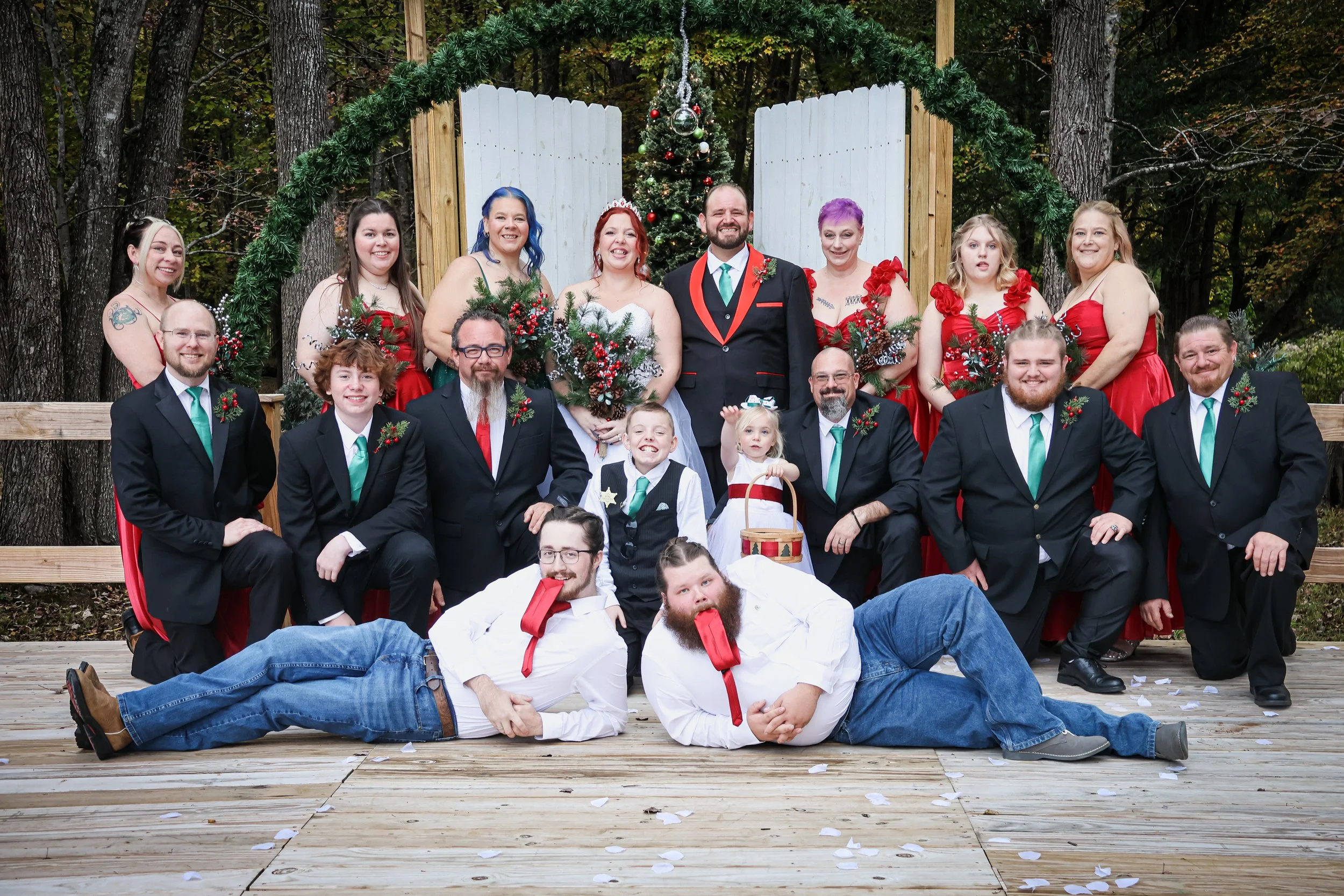 Group of people in formal and festive attire gathered outdoors in front of a decorated Christmas tree, with some sitting and others standing on a wooden platform surrounded by trees.
