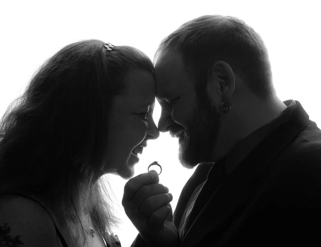 Silhouette of a smiling woman and a man touching foreheads, holding an engaged ring, with their eyes closed.