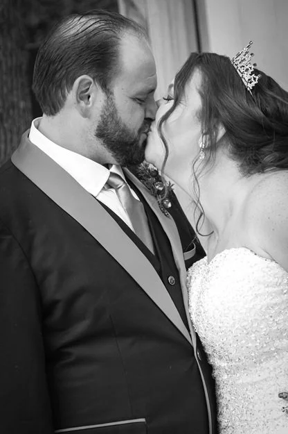 A black and white photo of a couple in wedding attire sharing a kiss, with the bride wearing a tiara and the groom in a suit with a tie.