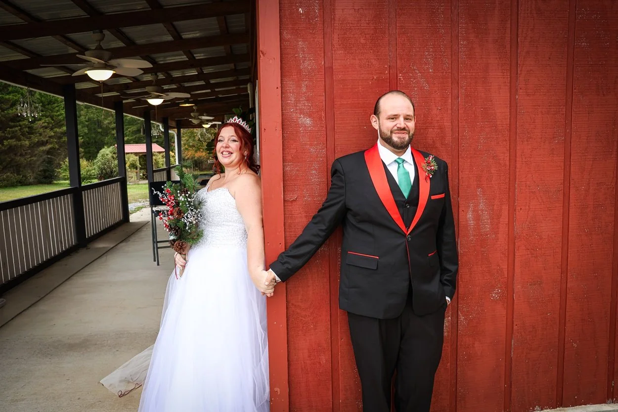 Bride and groom holding hands, standing back-to-back with the bride smiling and the groom smiling slightly, outdoors in a rustic setting with a red barn wall.