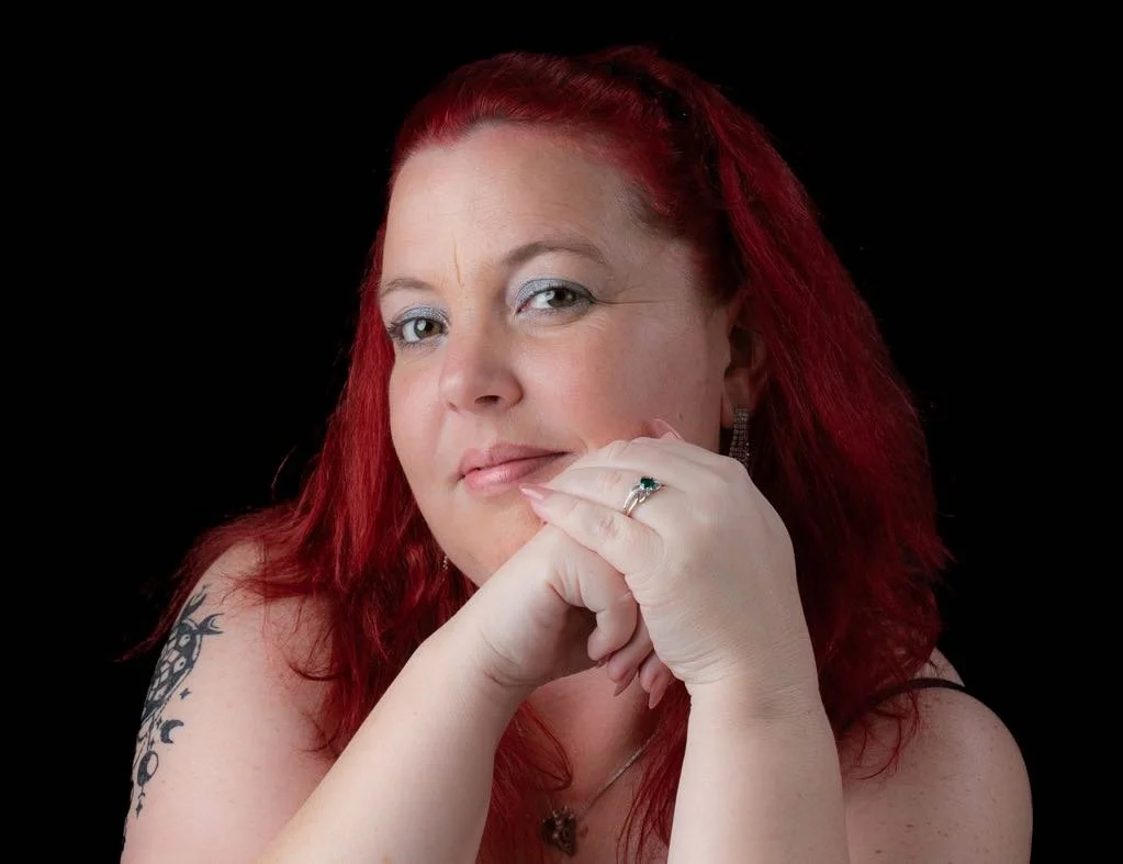 Portrait of a woman with red hair, wearing jewelry, resting her chin on her hand, against a black background.