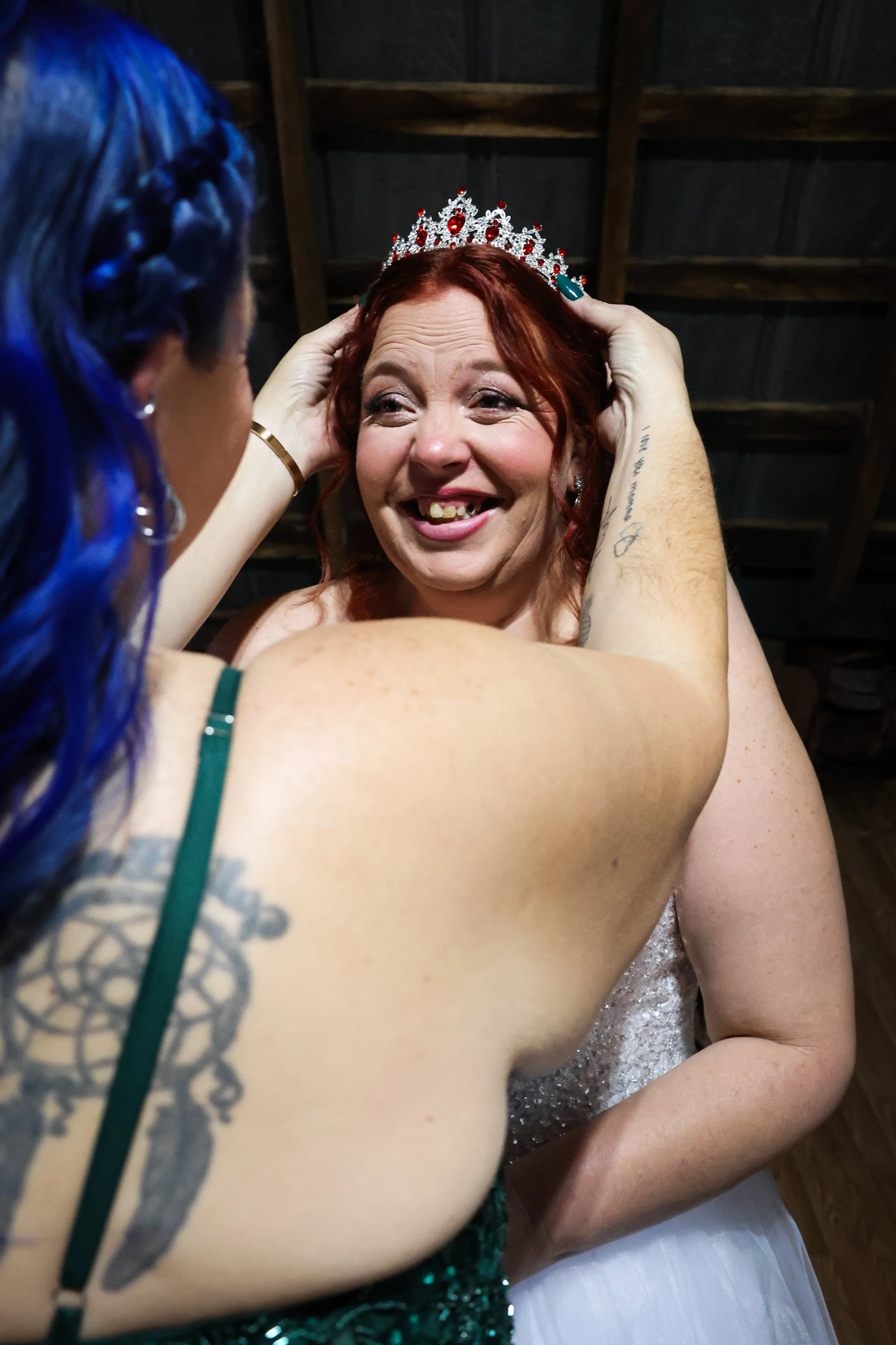 A woman with red hair and wearing a silver dress is being crowned with a sparkly tiara by another woman with blue braided hair. The woman with the tiara has a big smile and is looking up at the recipient.
