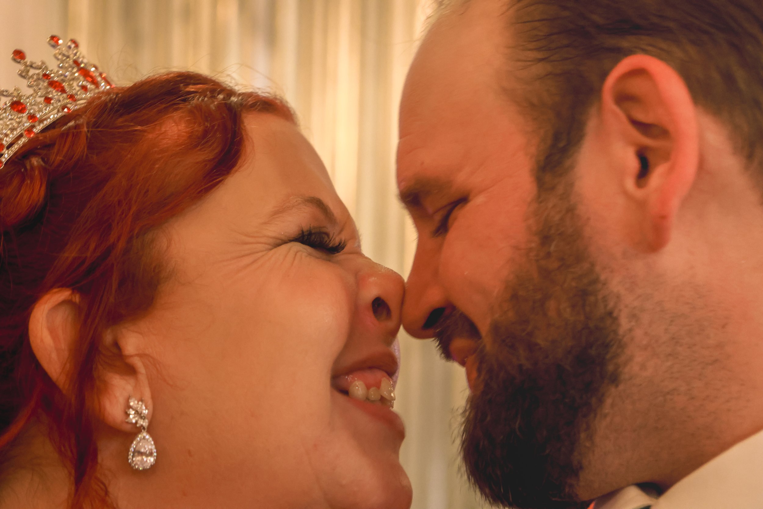 Close-up of a smiling bride and groom touching foreheads, holding hands, at their wedding. The bride is wearing a tiara and earrings.