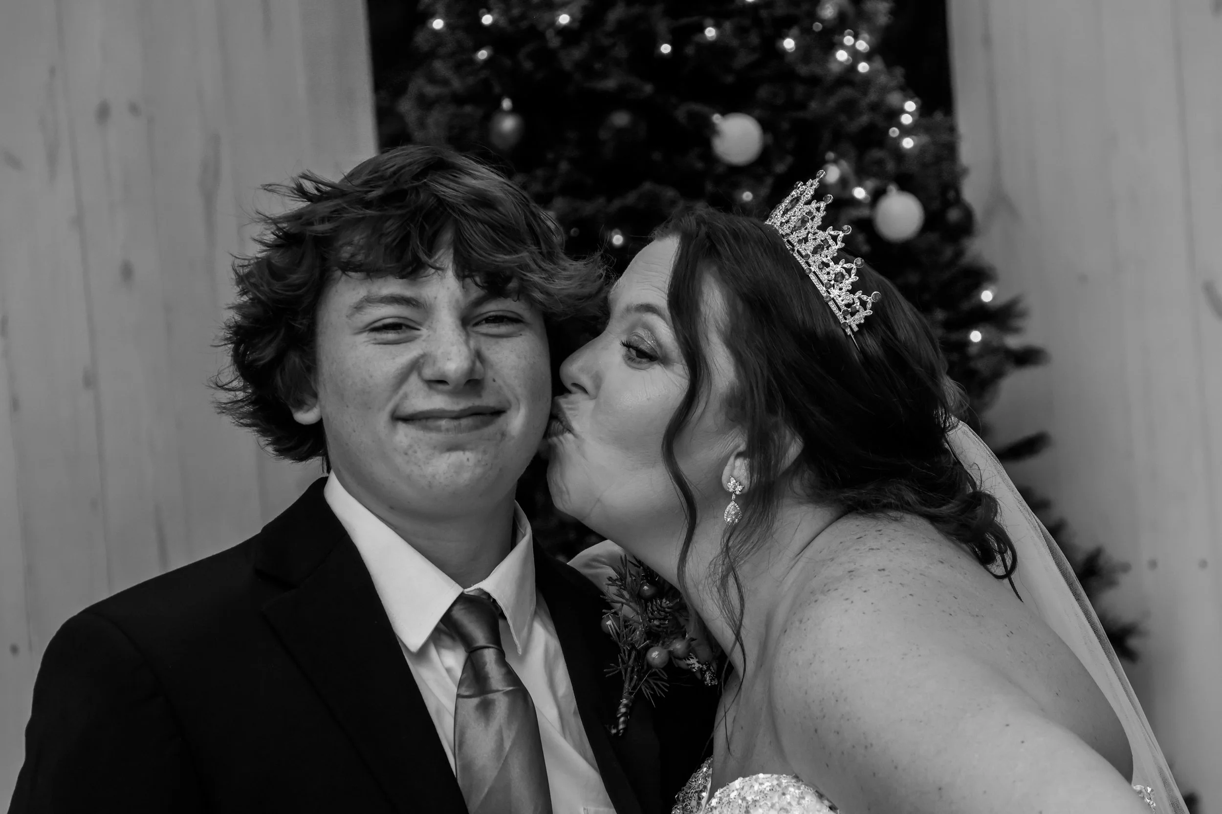A black-and-white photo of a couple at a wedding, with the woman kissing the man's cheek. The woman is wearing a tiara and earrings, and the man is dressed in a tuxedo with a bow tie. There is a decorated Christmas tree in the background.