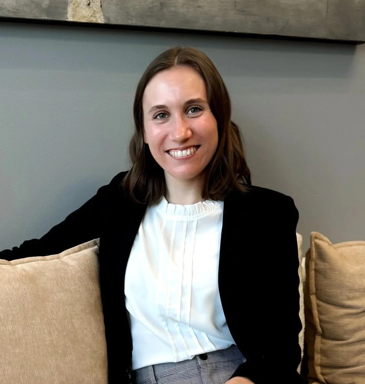 Laura Esposito. Therapist (LCPC licensed) with shoulder-length brown hair smiling, wearing a white blouse and black jacket, sitting on a beige couch inside a modern room.