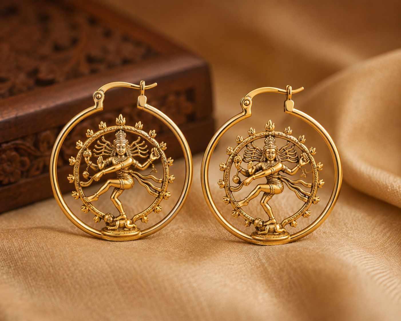 Gold earrings with intricate designs of a dancing deity in a circular frame, displayed on a beige fabric surface with a carved wooden box in the background.