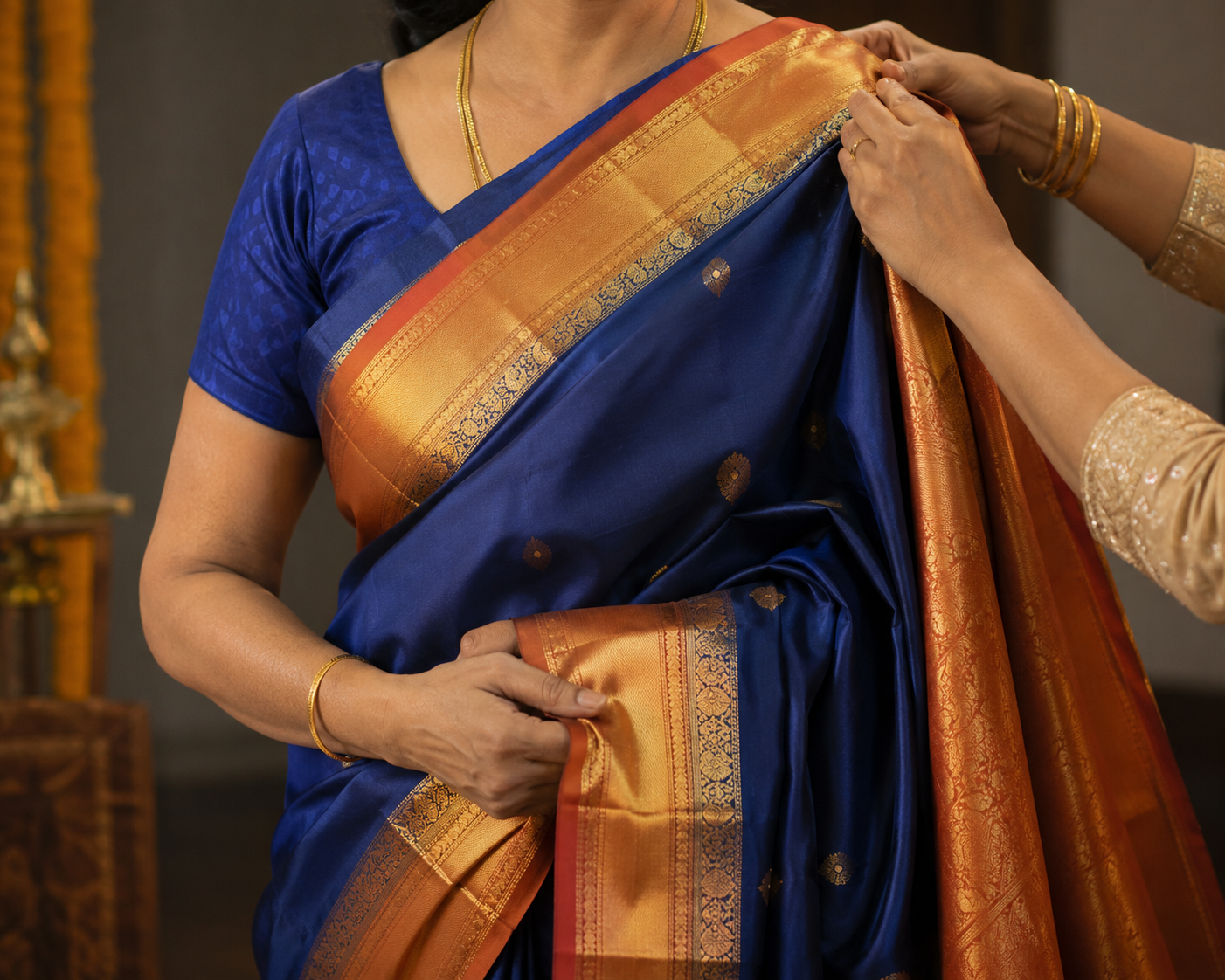 A woman wearing a traditional blue and orange sari, being help drape the sari with gold jewelry visible, including a necklace and bangles.