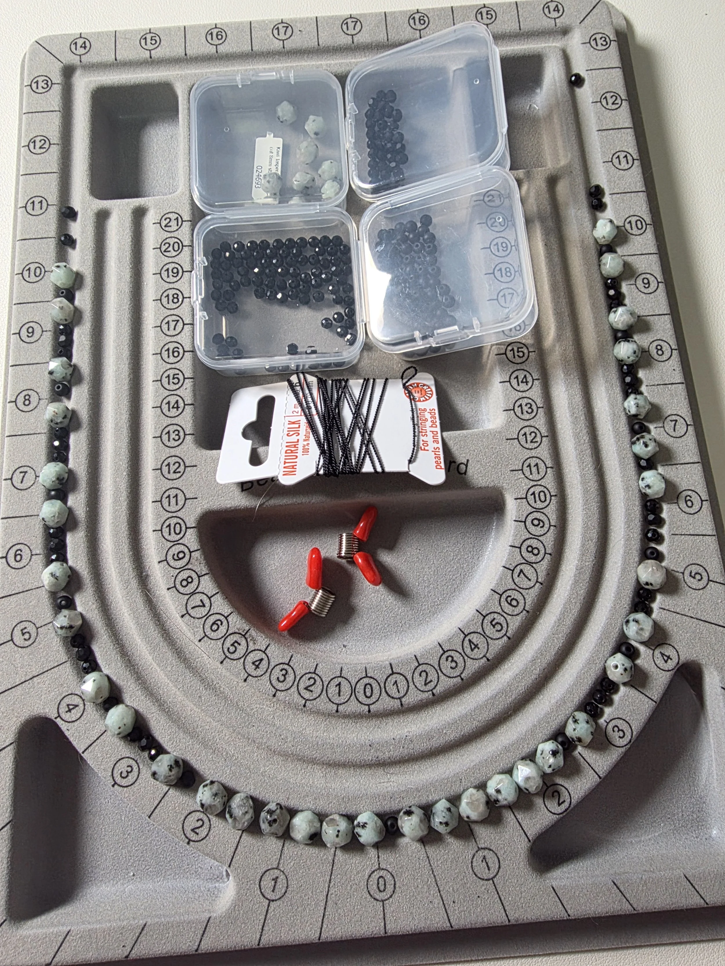 A beading mat with numbered rings and a spiral pattern, three small plastic containers filled with black, white, and gray beads, a pack of black headpins, and several red and silver earring hooks.