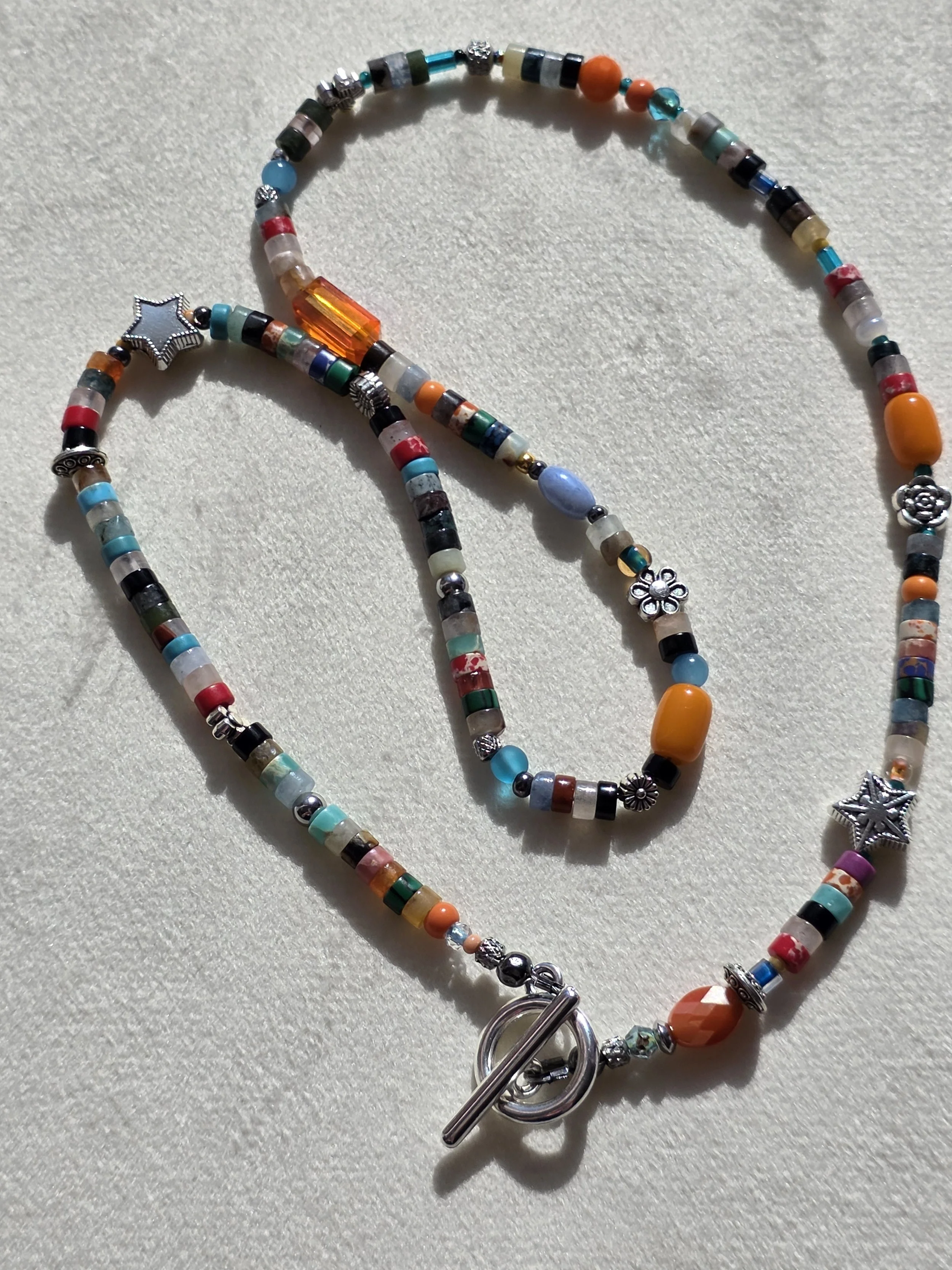 Handmade beaded necklace with mixed gemstone heishi beads in turquoise, carnelian and jasper tones, amber glass beads, and silver star and flower spacers, laid flat on a neutral background