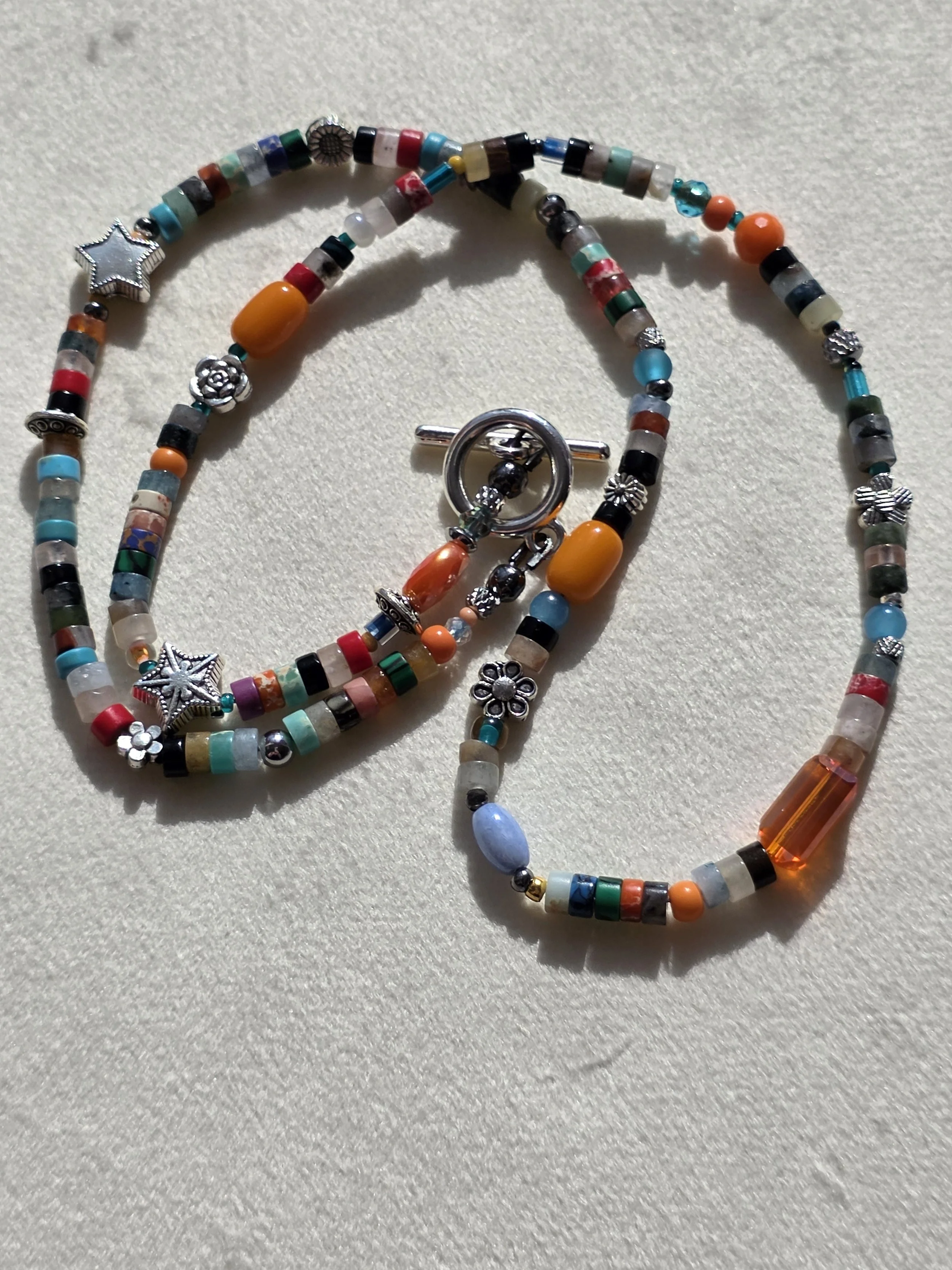Handmade beaded necklace with mixed gemstone heishi beads in turquoise, carnelian and jasper tones, amber glass beads, and silver star and flower spacers, laid flat on a neutral background