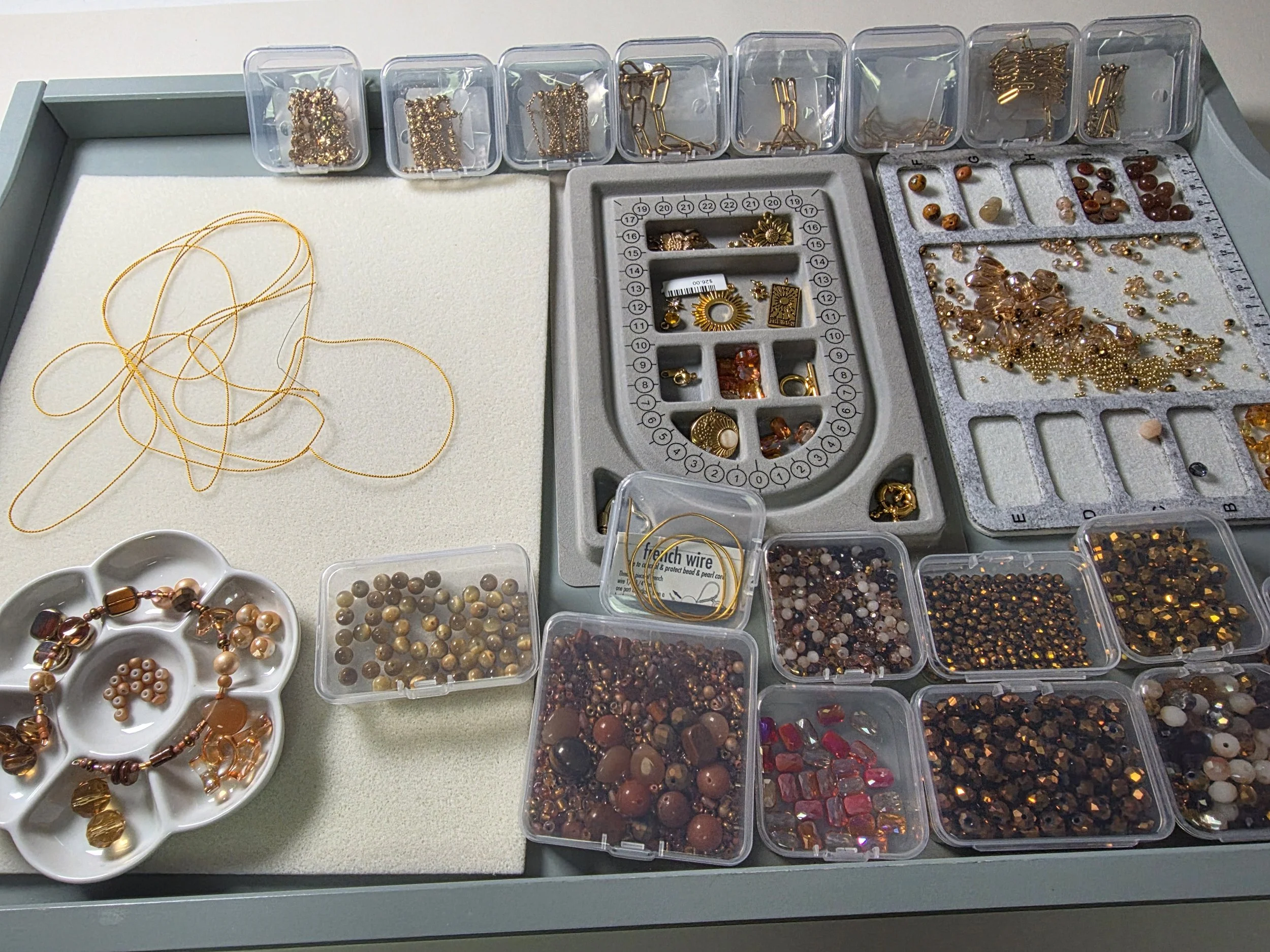 A jewelry-making station with various containers holding beads, chains, and jewelry components, along with some finished jewelry pieces on a white tray and small dish.