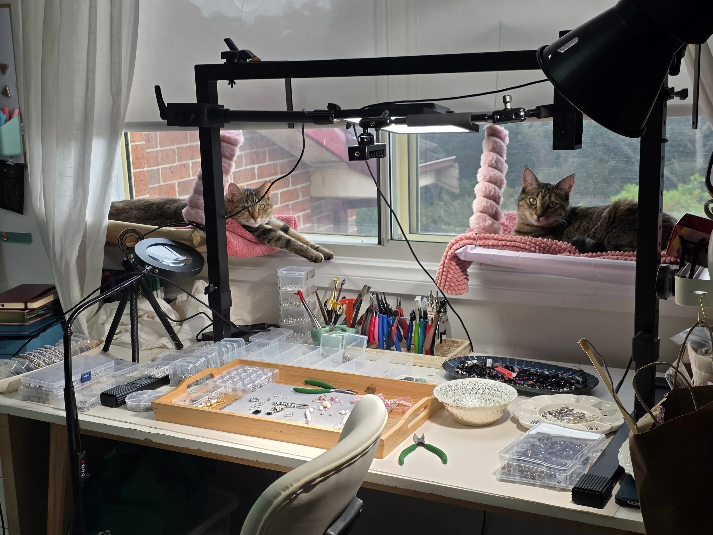 A cluttered workspace with two cats resting on a windowsill, surrounded by crafting or jewelry-making supplies, tools, and boxes.