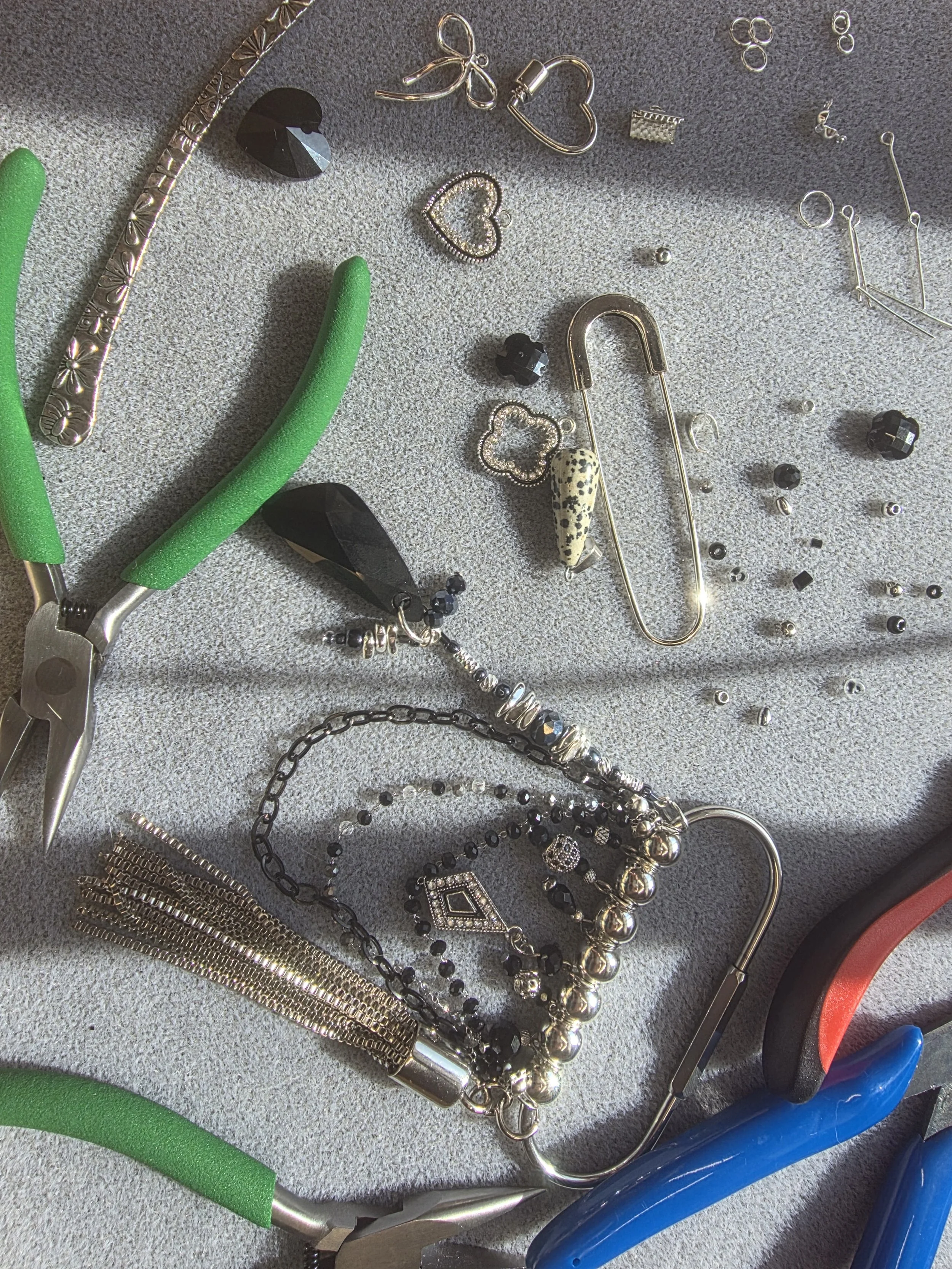A collection of jewelry and tools spread out on a surface. The jewelry includes various necklaces, rings, earrings, and a safety pin with decorative elements. The tools include pliers with green handles and a blue-handled tool, likely used for jewelry assembly or repair.