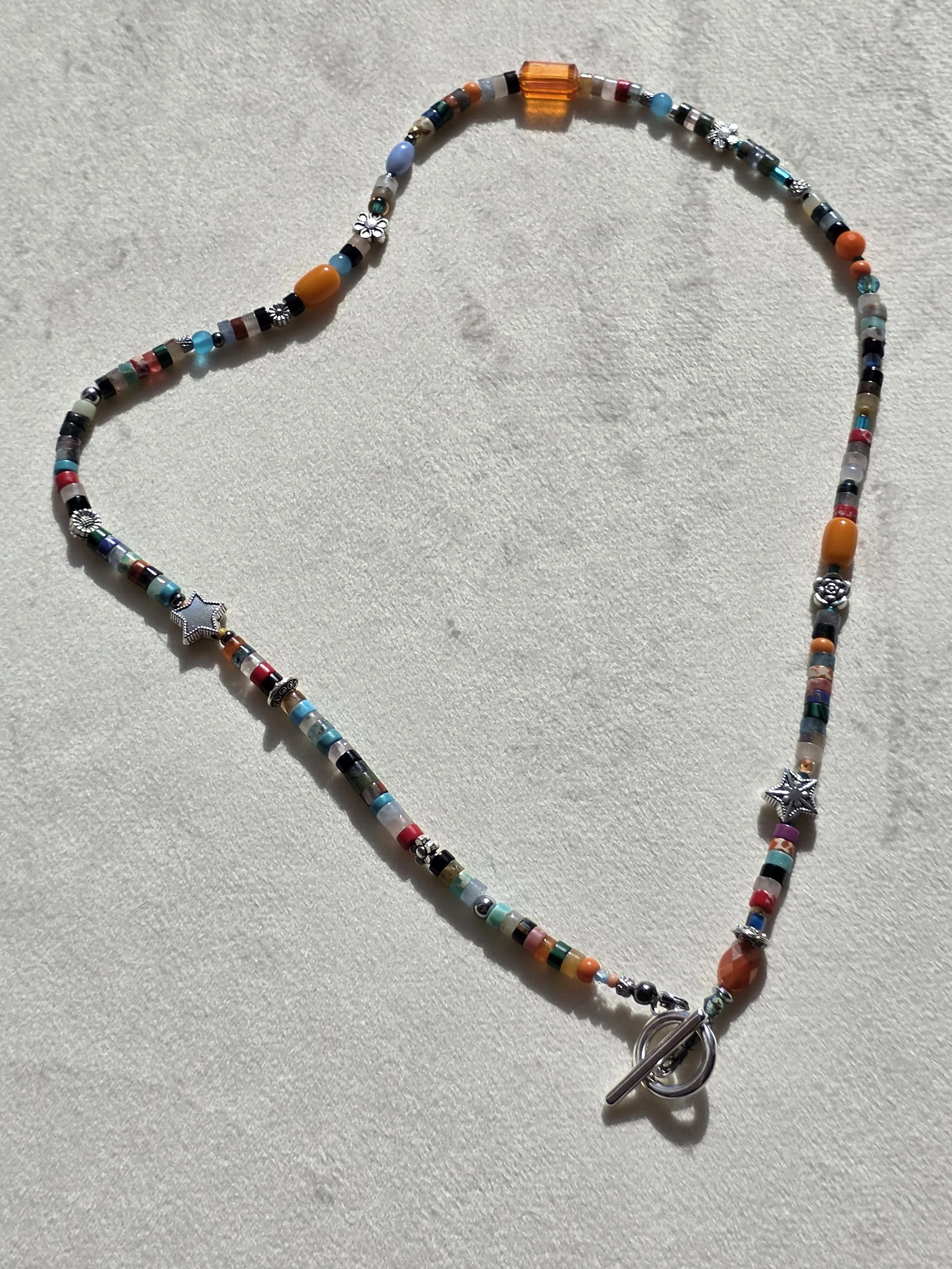 Handmade beaded necklace with mixed gemstone heishi beads in turquoise, carnelian and jasper tones, amber glass beads, and silver star and flower spacers, laid flat on a neutral background