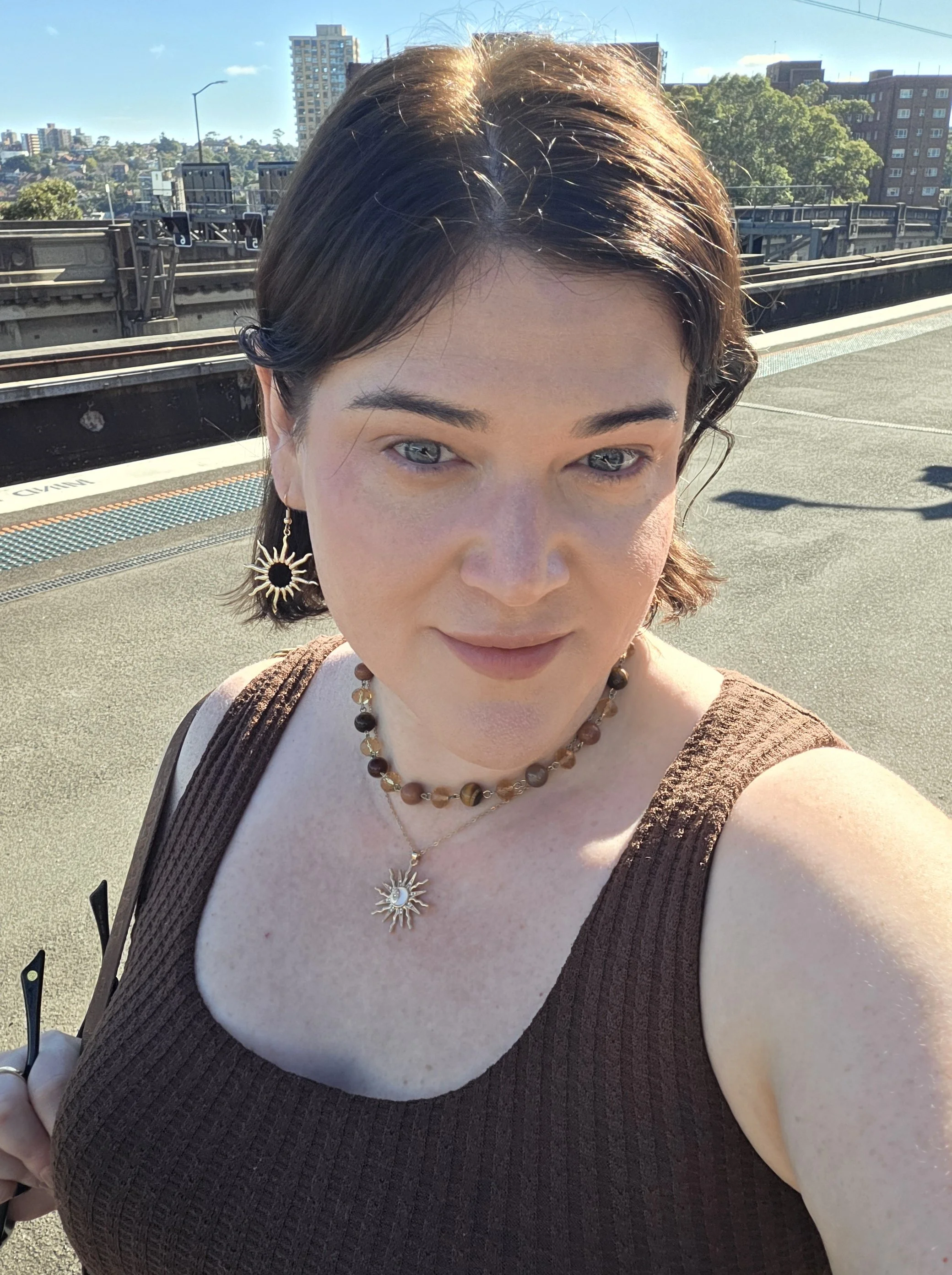 A woman with short dark hair and light blue eyes taking a selfie outdoors on a city street during daytime. She is wearing jewelry including sun-shaped earrings and layered necklaces. The background shows buildings, trees, and part of a railway or highway.