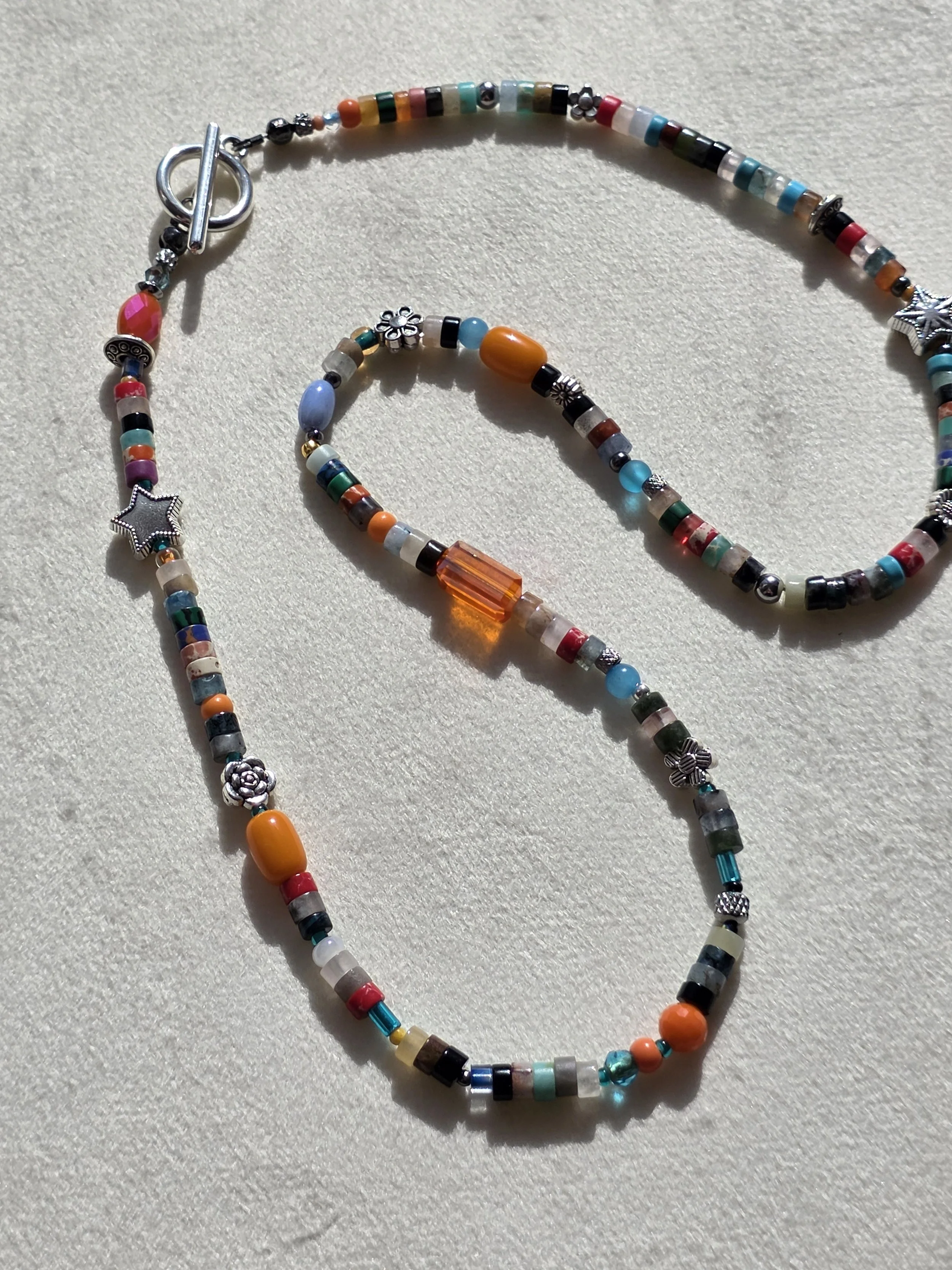 Handmade beaded necklace with mixed gemstone heishi beads in turquoise, carnelian and jasper tones, amber glass beads, and silver star and flower spacers, laid flat on a neutral background