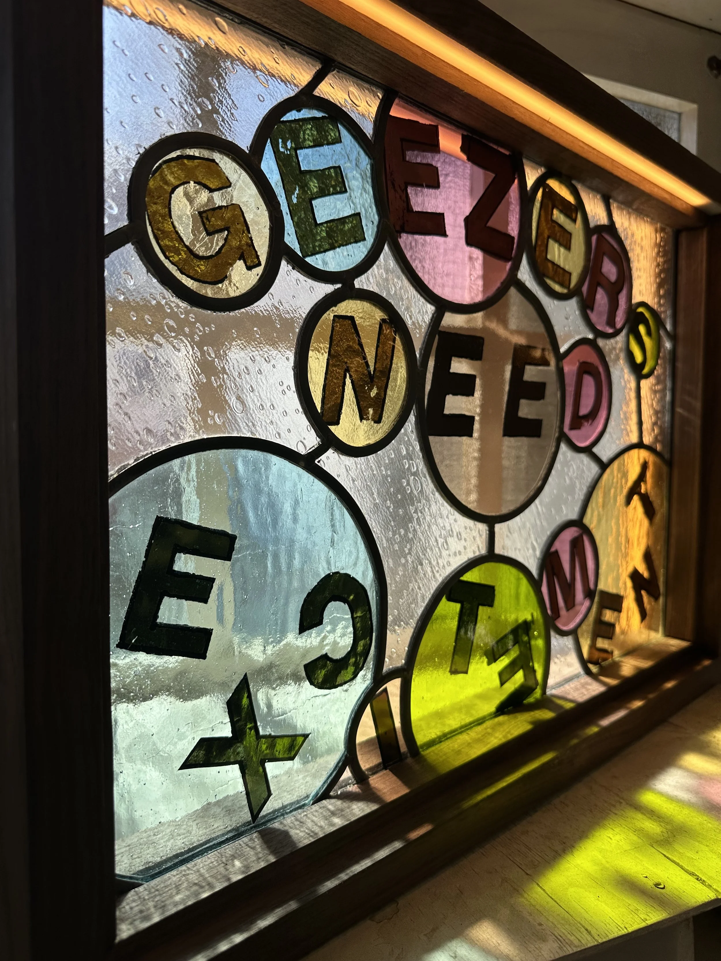 Colorful stained glass window with the word 'GEEZER' spelled out in individual letters, with raindrops on the glass and a wooden frame surrounding it.