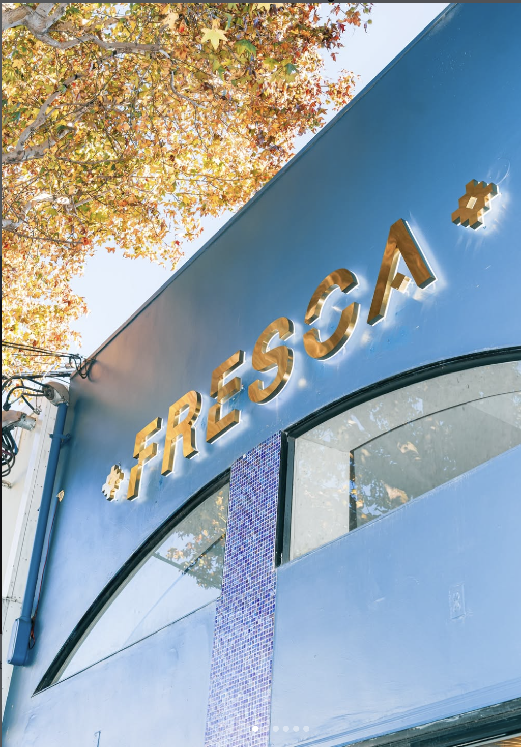 Close-up of a blue storefront with golden 3D letters spelling 'FRESCA' and a small pixelated star logo, with autumn leaves on a tree above.