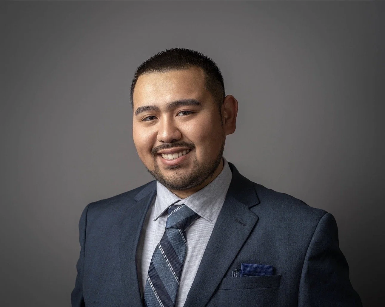 Professional headshot of a smiling man wearing a dark gray suit, light blue shirt, and striped tie, with a pocket square, against a gray background.