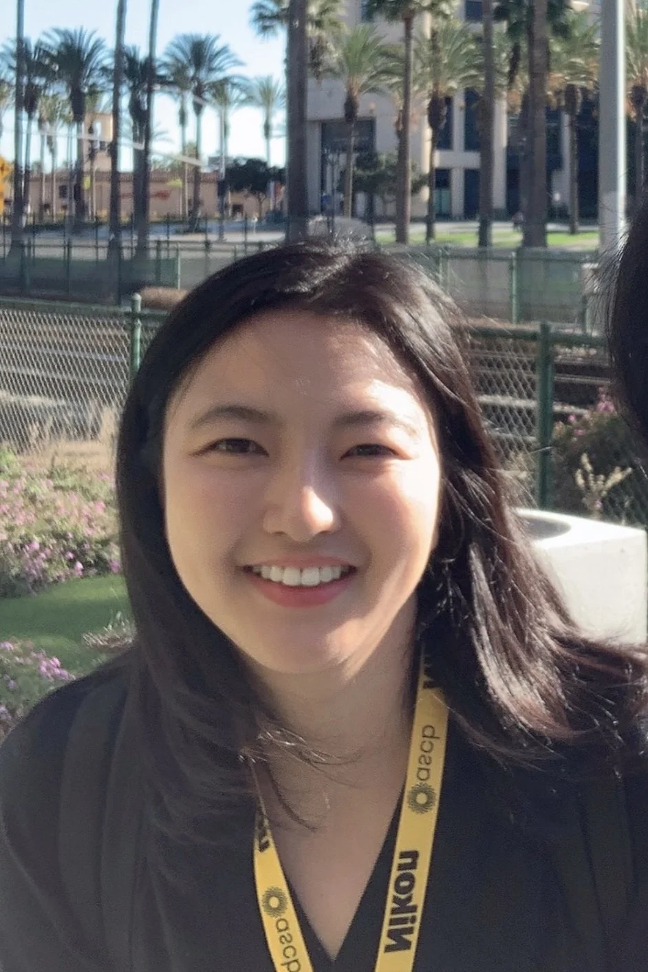 A woman smiling outdoors with palm trees and buildings in the background, wearing a black shirt and a yellow ID badge that says Nikon and decp.