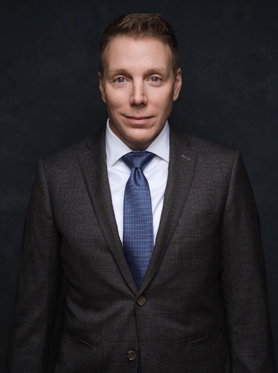 Cameron, a friendly, professional man with short blond hair, blue eyes, and fair skin wearing a dark suit, white shirt, and blue tie, standing against a black background.
