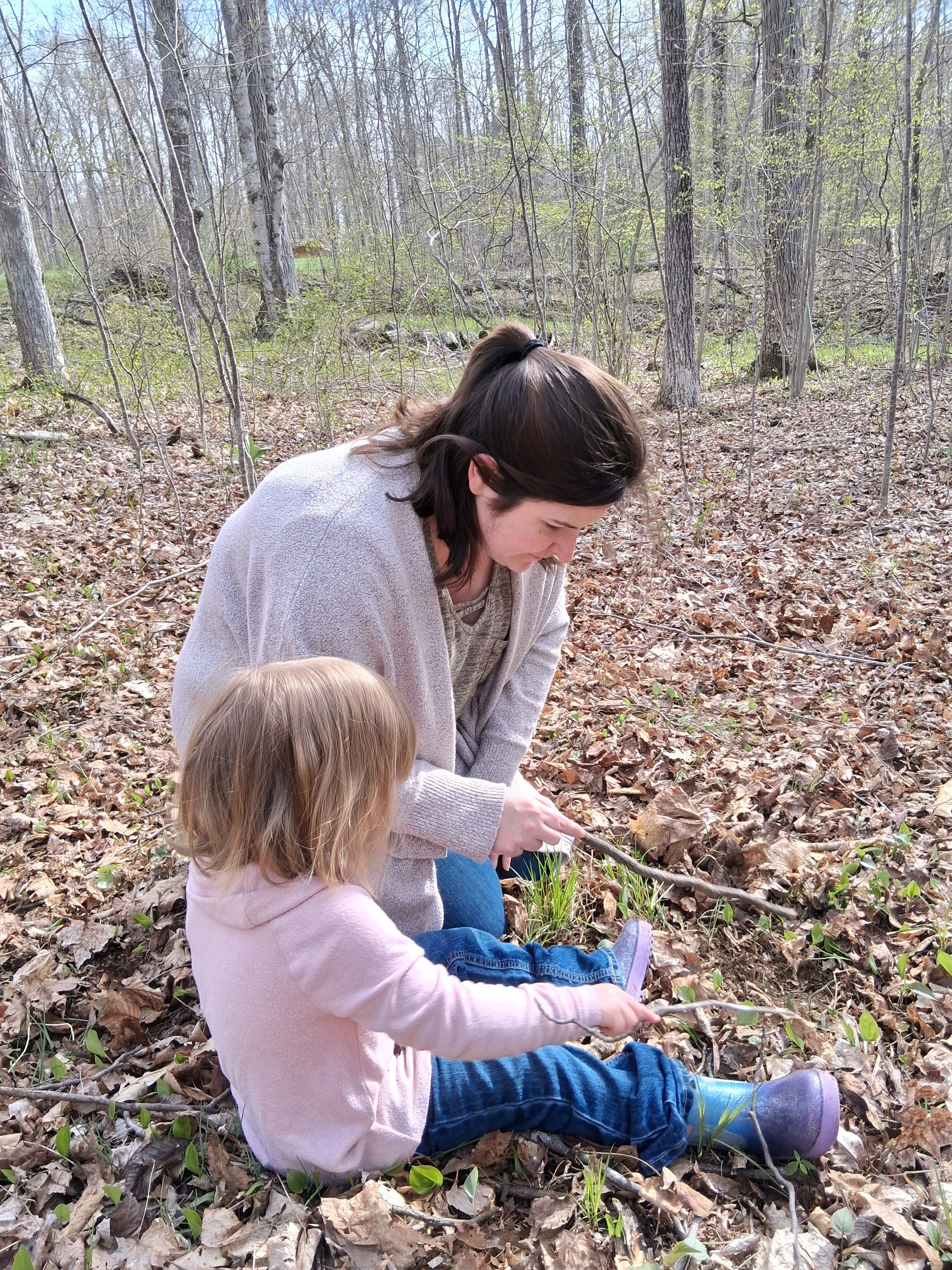 Child guide working alongside child to move leaves with sticks. A quiet moment in a natural preschool.