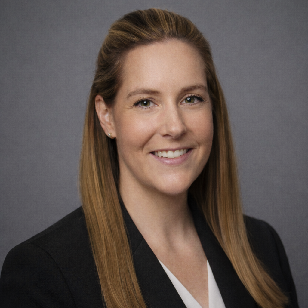 A professional headshot of a smiling woman with long straight red hair, wearing a black blazer and white top, against a gray background.