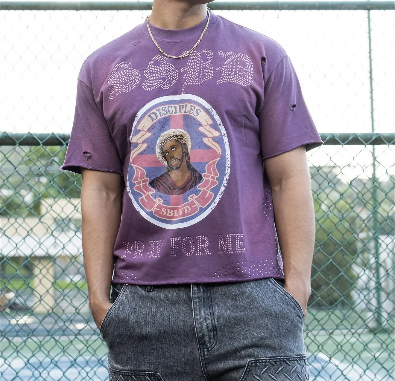 Person wearing a purple t-shirt with a graphic of Jesus Christ with a halo, surrounded by text that reads 'Disciples SBVD' and 'Pray for Me', standing outdoors in front of a chain-link fence.