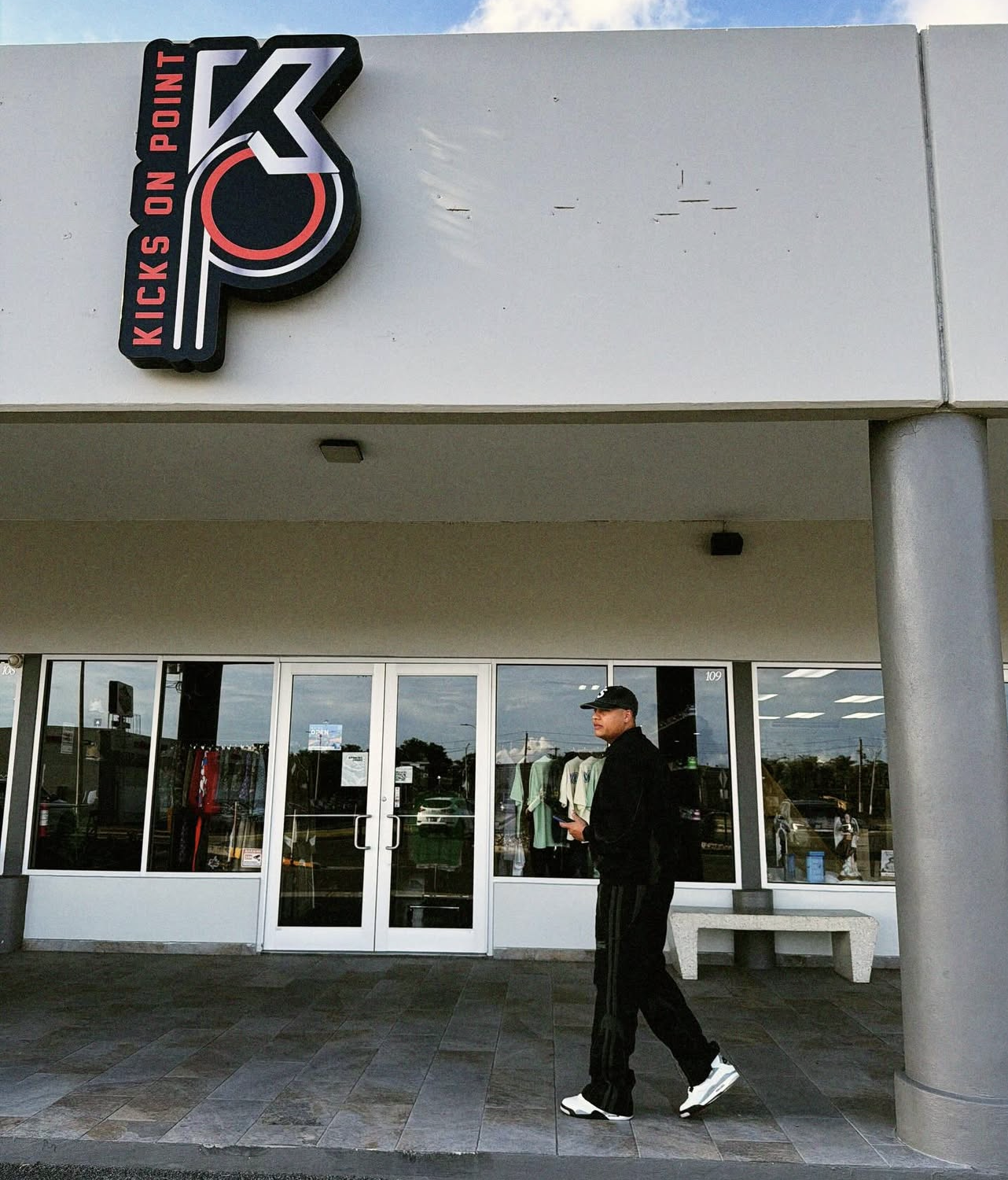 Storefront with a sign that reads "Kicks on Point" and a logo, with a person in black athletic clothing and a cap walking in front of the entrance.