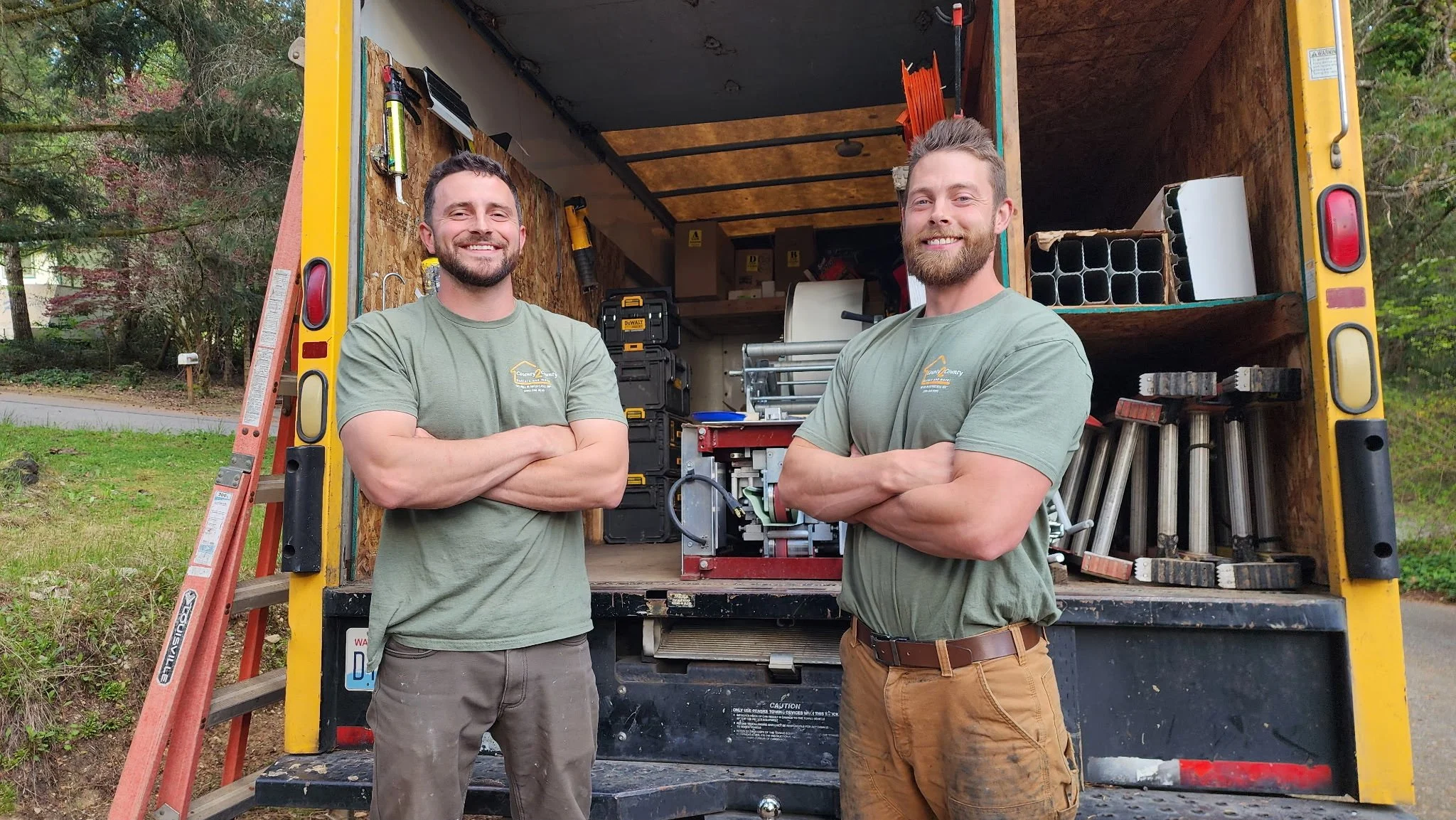Two men standing in front of a yellow work truck with tools inside, smiling with crossed arms, outdoors with trees in the background.
